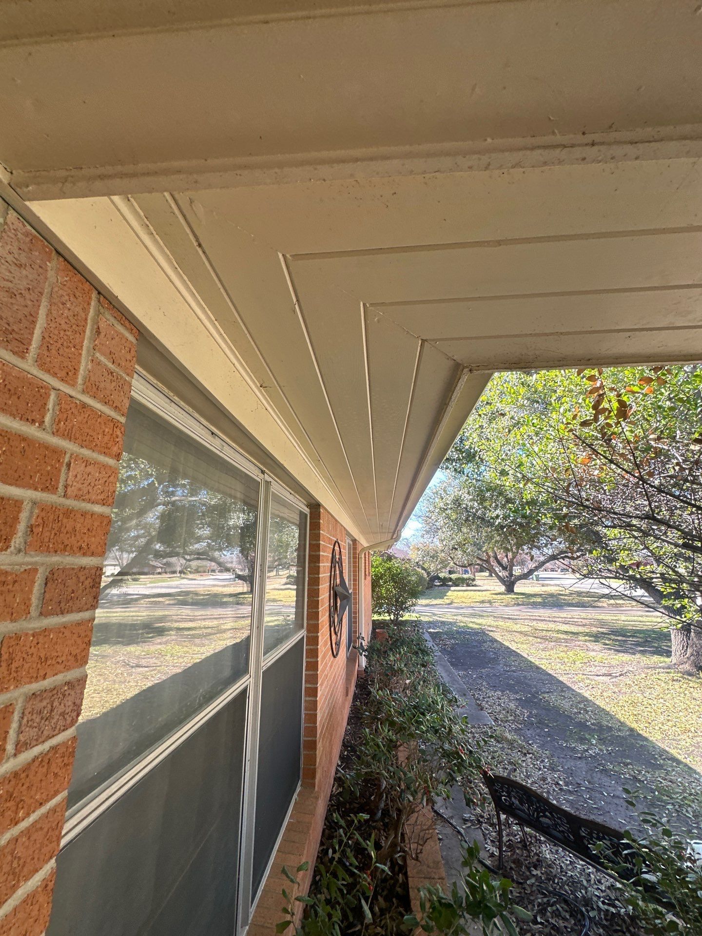 Brick house side porch with white eaves, screen window, and trees in a sunny yard