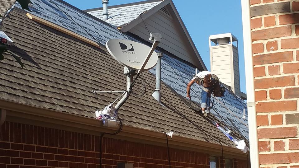 Person on a roof installing or adjusting a satellite dish beside a brick house.