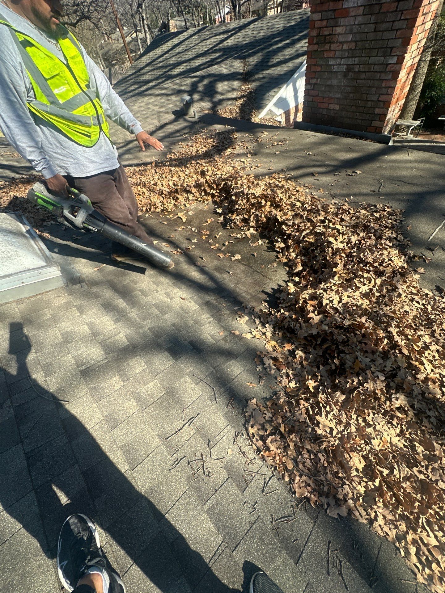 Worker in a reflective vest blowing leaves from a driveway with a leaf blower near a brick house and trees.