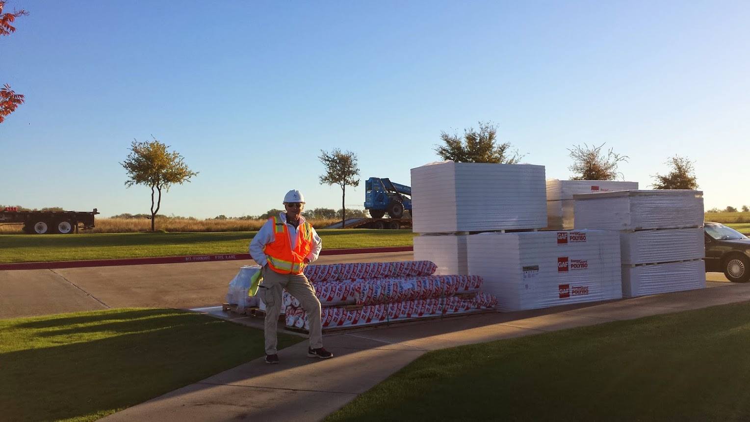 Worker in safety vest standing beside stacked boxes and pallets outdoors at sunset.