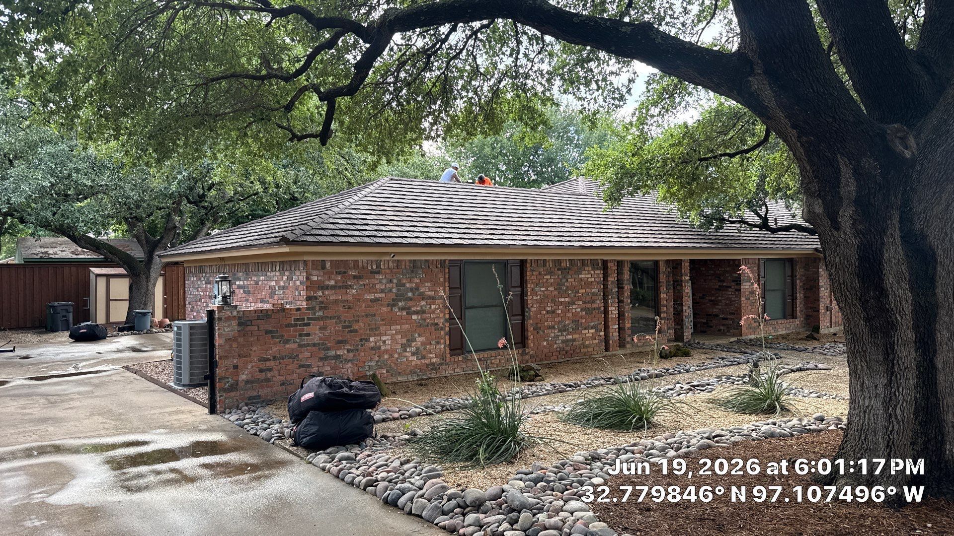 Brick house with a damaged roof and a black trash bag in the driveway, under large trees