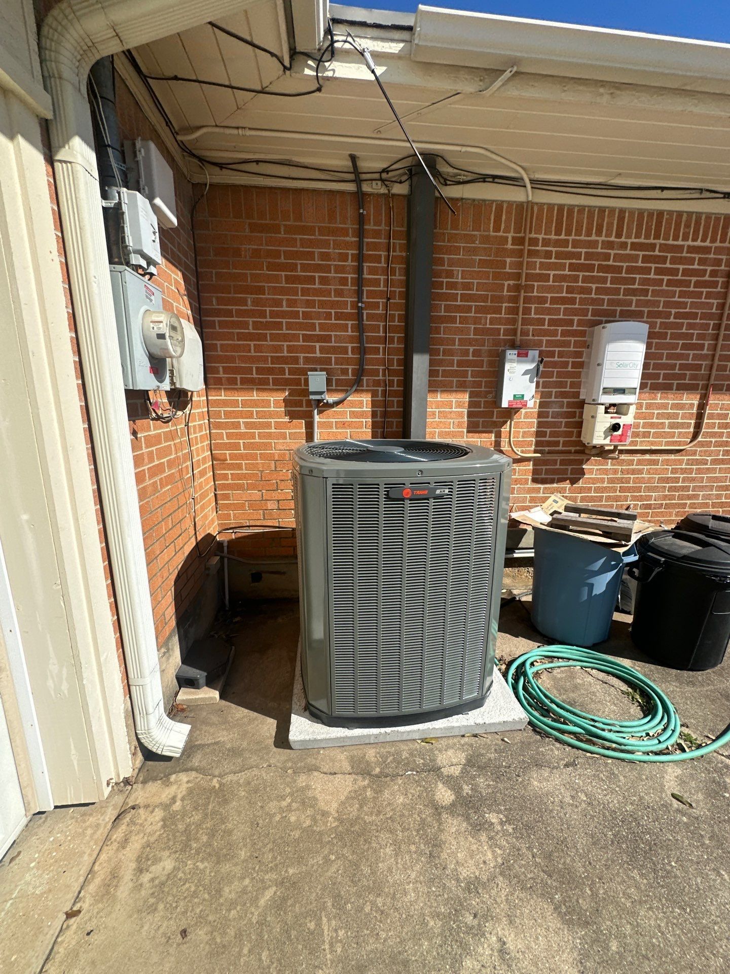 Outdoor HVAC unit beside a brick wall, with coiled green hose and trash bins on a concrete patio.
