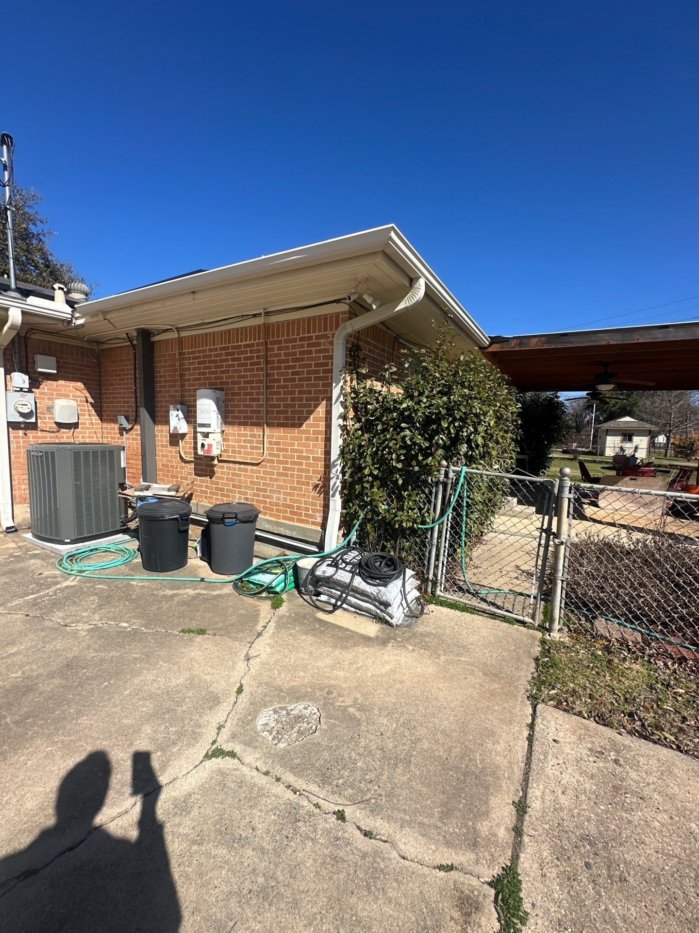 Brick house exterior with driveway, trash bins, and covered carport under a clear blue sky