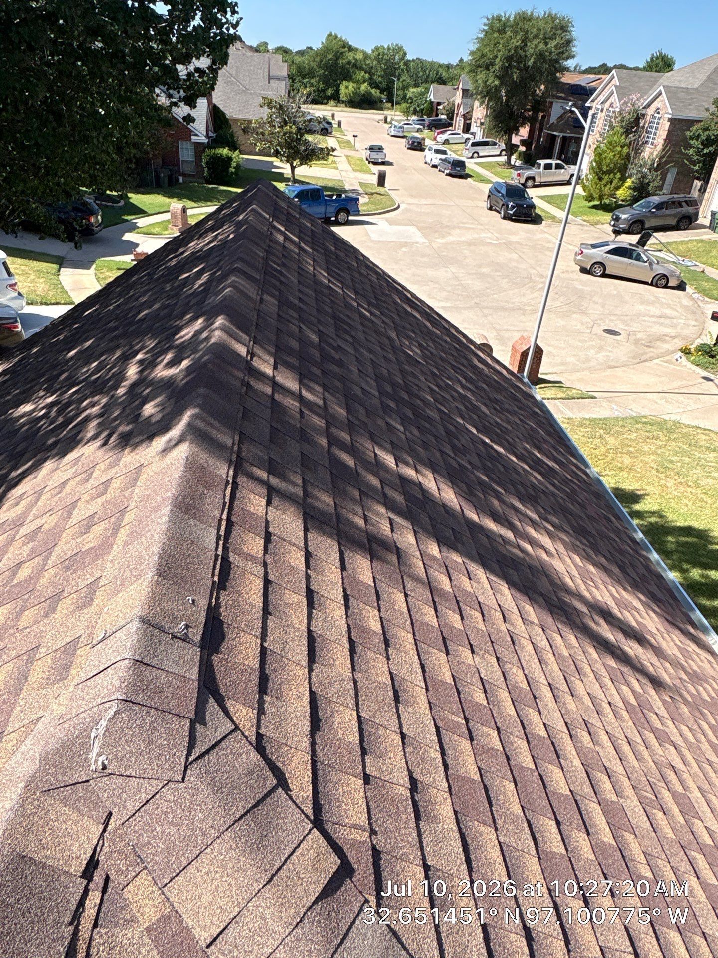View from a rooftop over a suburban street with houses, parked cars, and trees on a sunny day