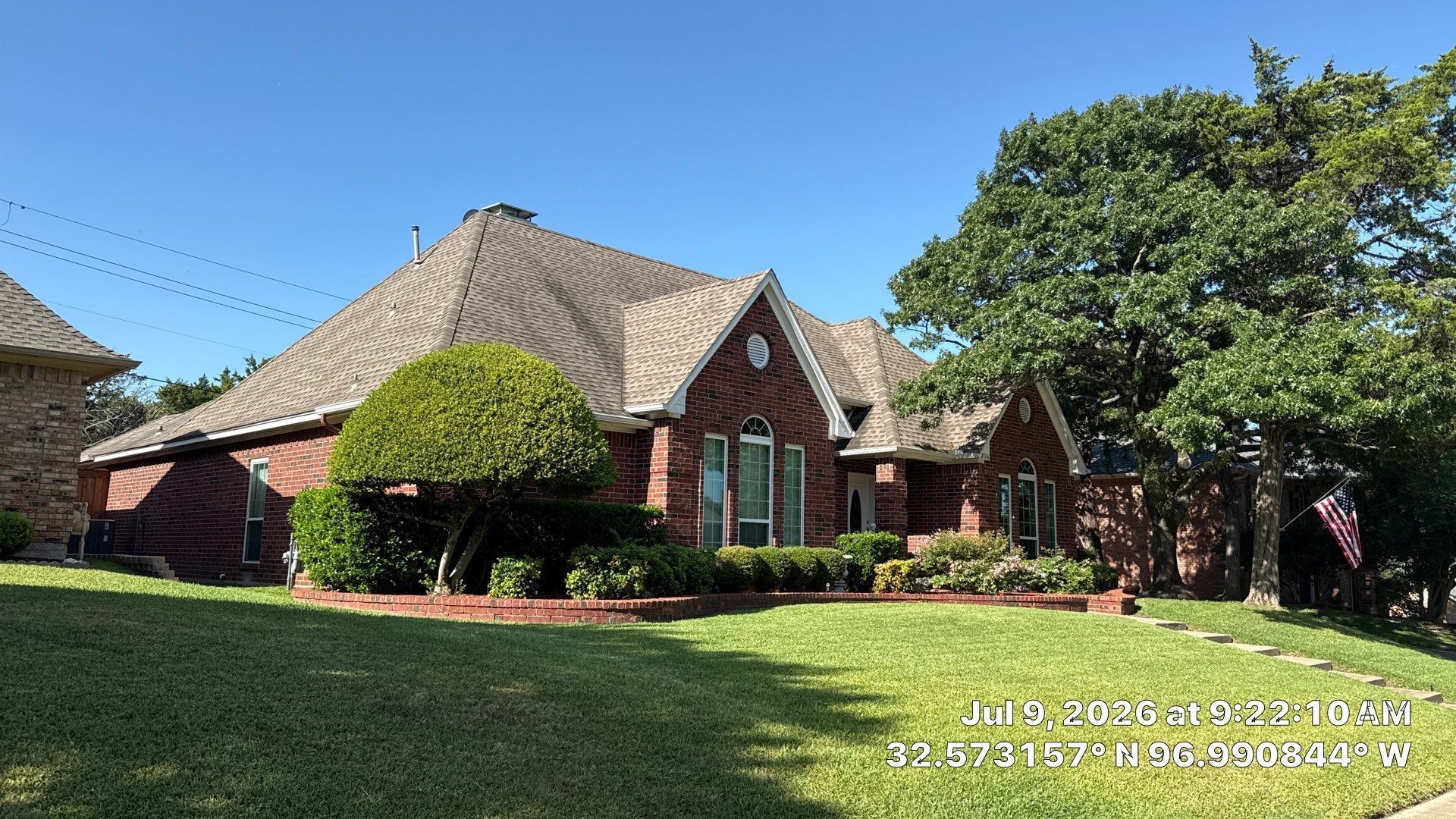 Brick suburban house with a steep roof, manicured lawn, and large trees under a clear blue sky