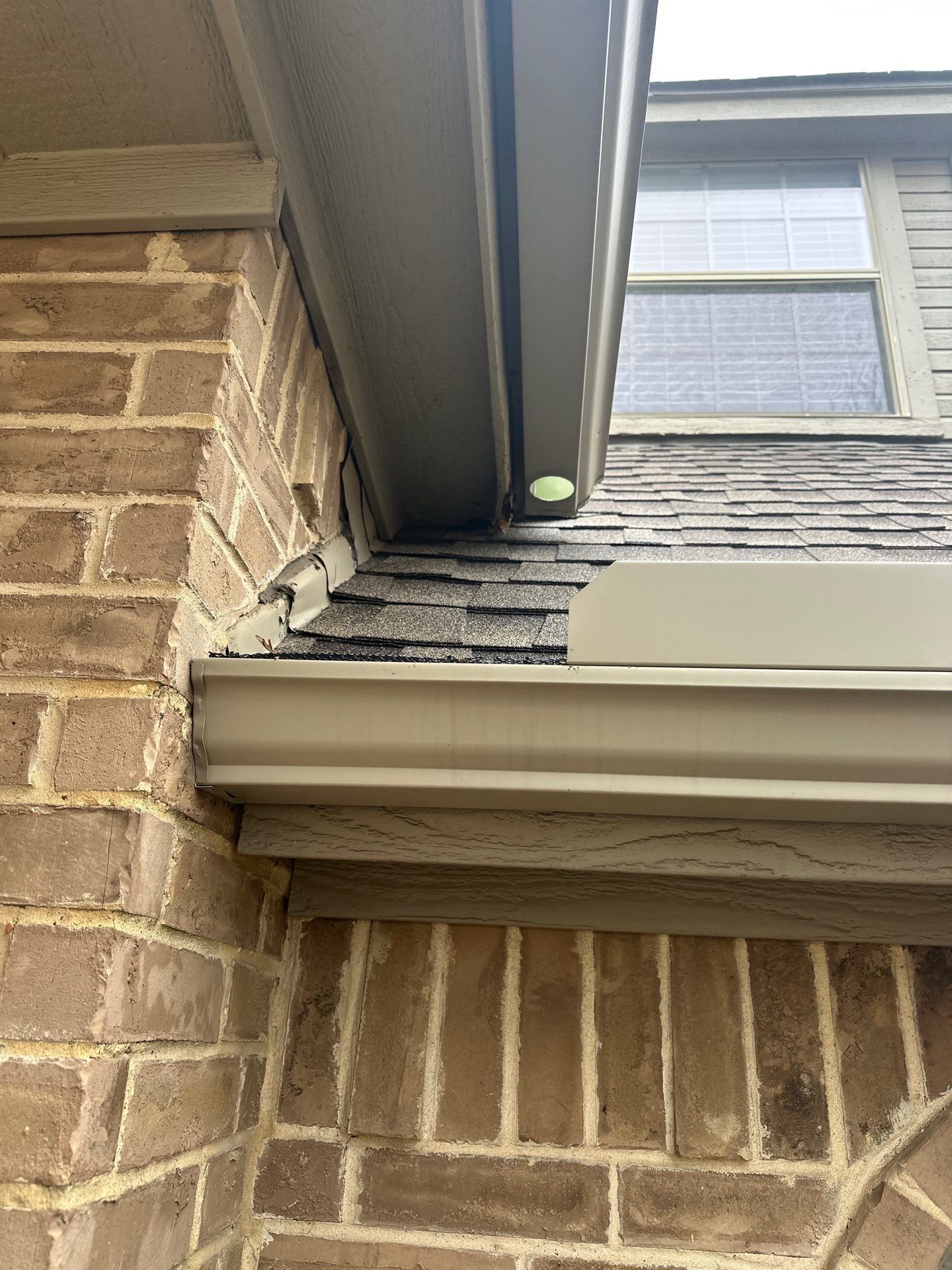 Close-up of a brick house exterior with roof eaves, gutter, and a window above a shingled roof edge