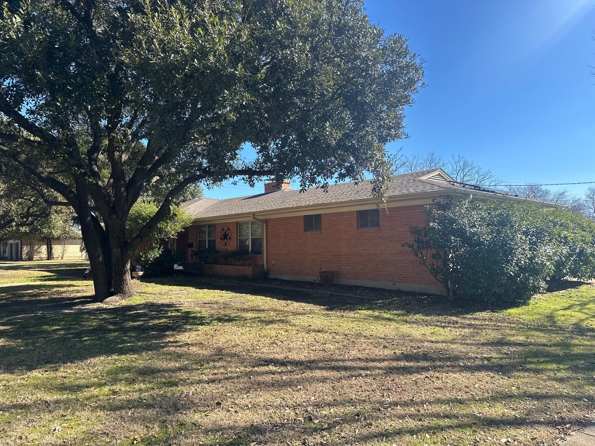 Single-story brick house with a large tree in the yard under a bright blue sky.