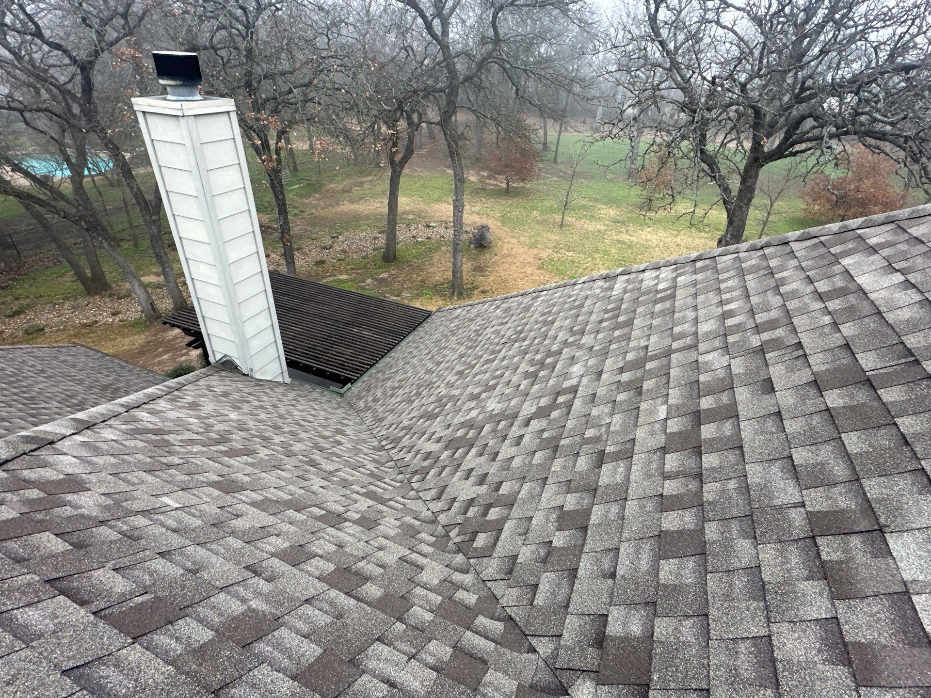 Gray shingled roof with a white chimney, overlooking leafless trees and a grassy yard