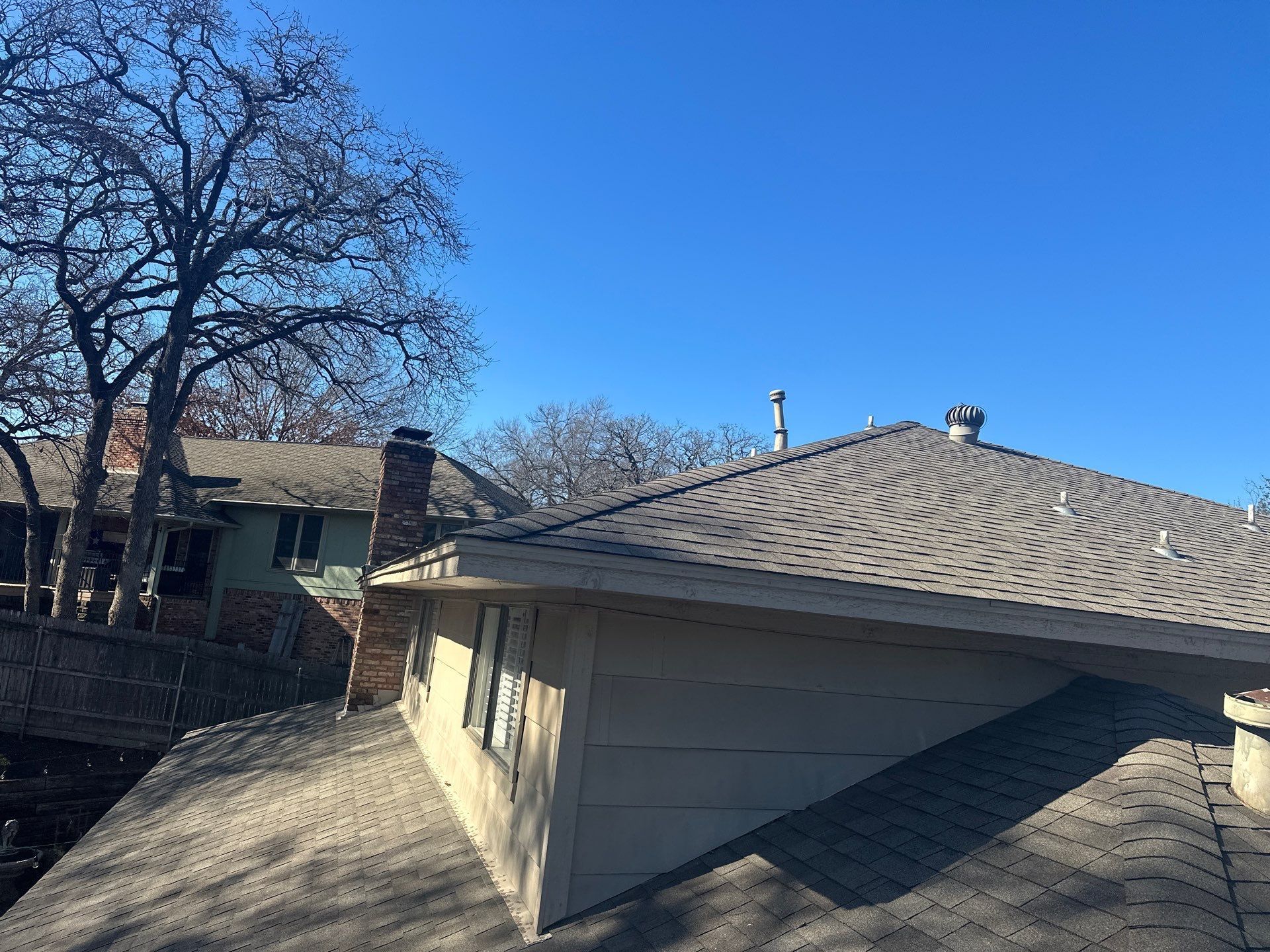Residential rooftops with gray shingles and bare trees under a bright blue sky
