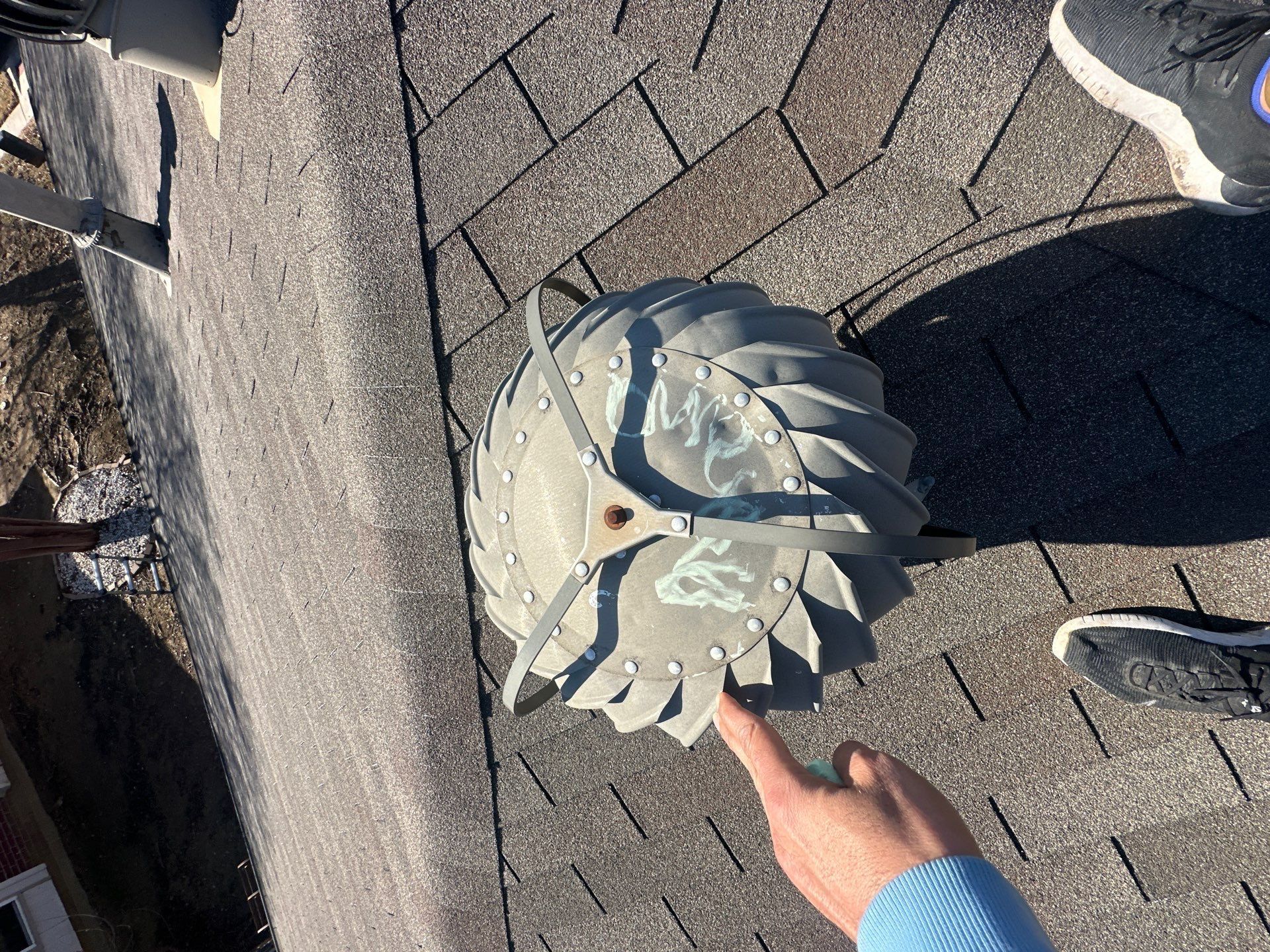 Hand pointing at a gray, flower-shaped roof vent on asphalt shingles near a curb