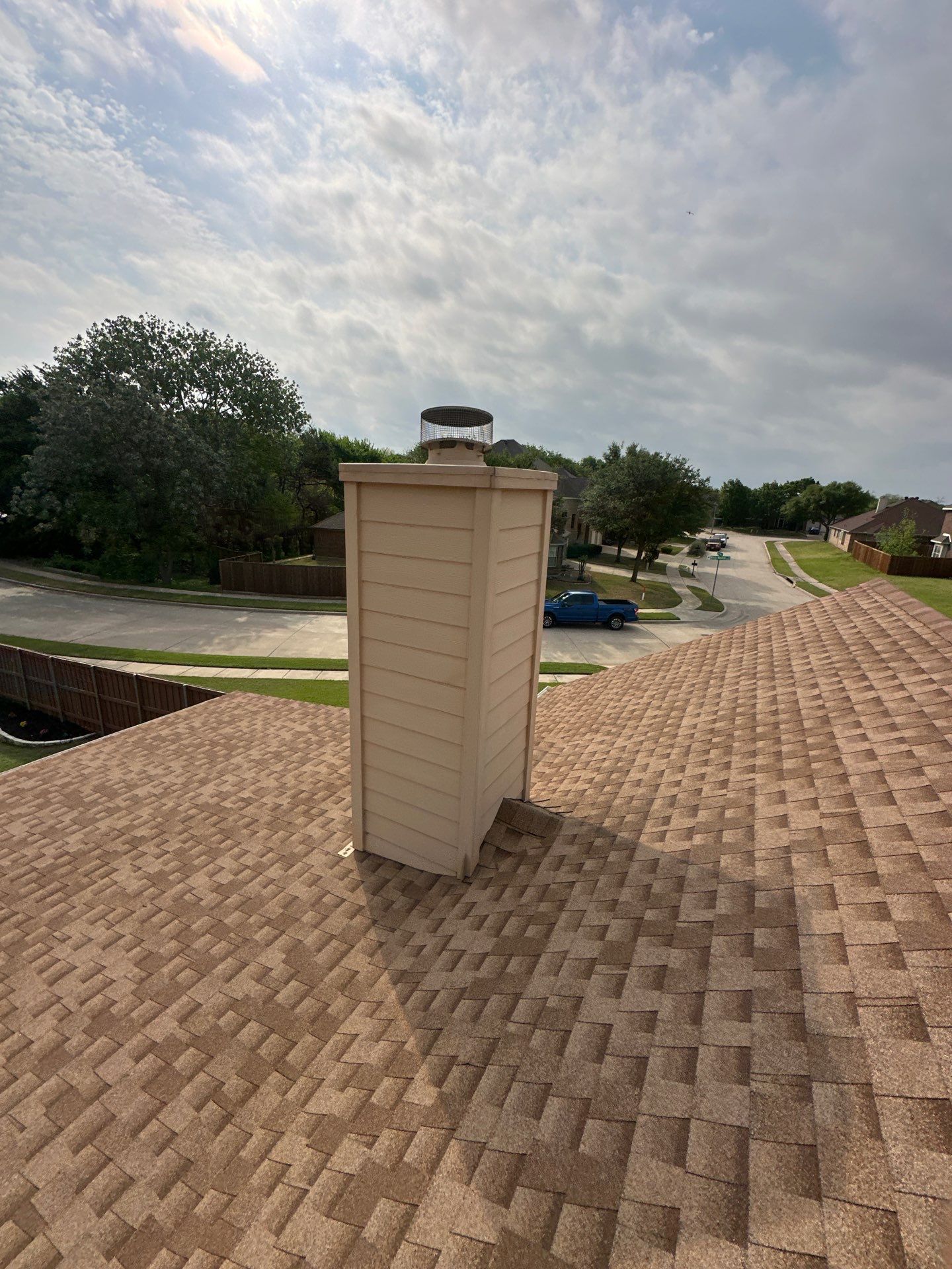 Brick chimney on a shingled roof under a cloudy sky, with trees and a street in the background.