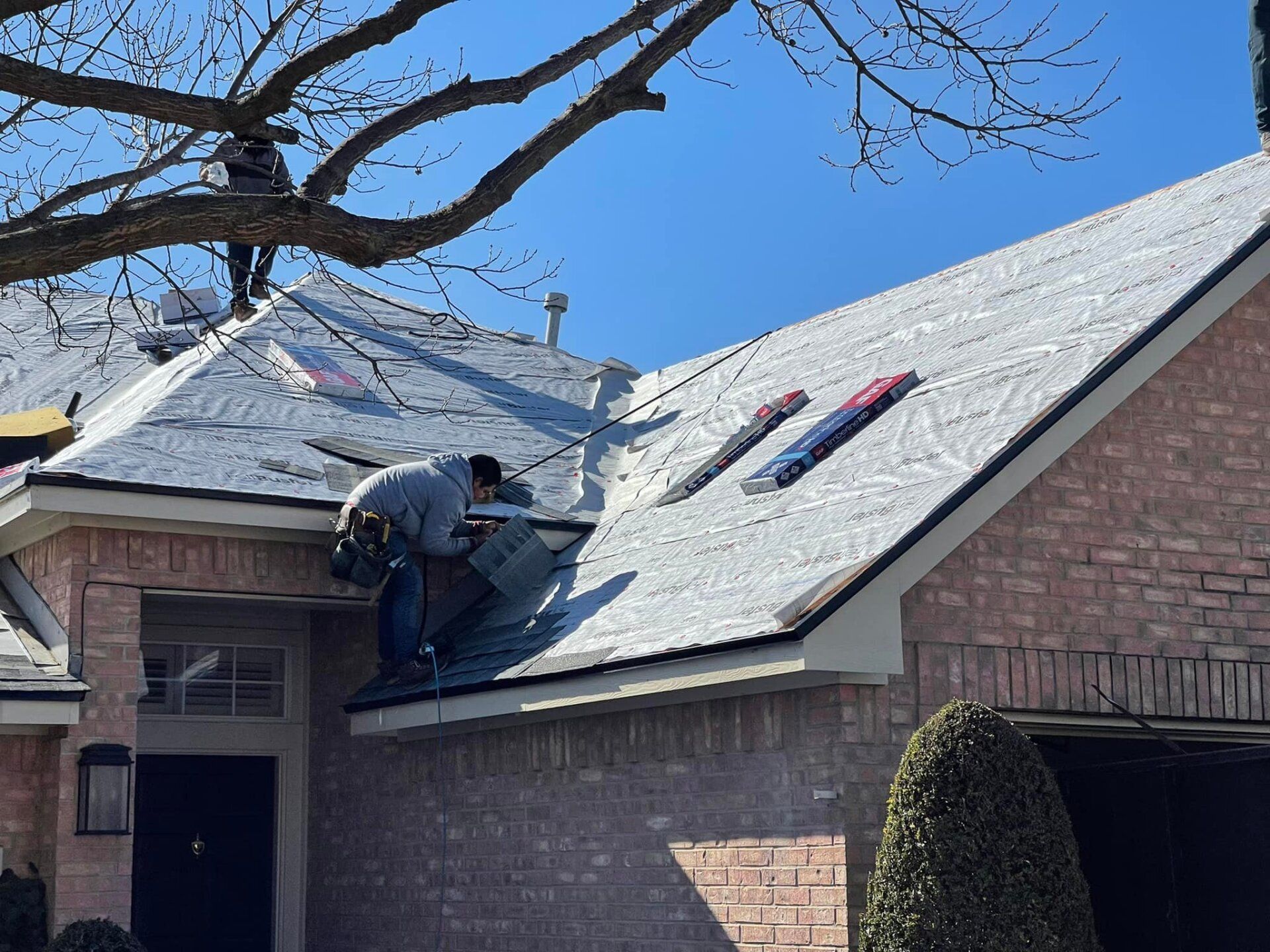 Workers repairing a shingled roof on a brick house under a blue sky