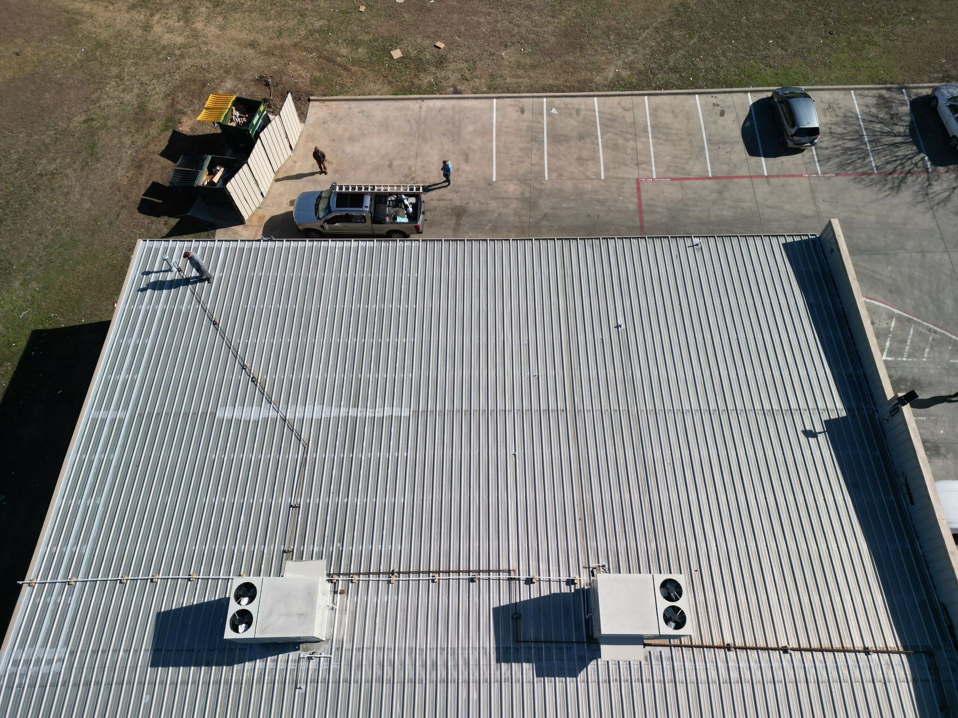 Aerial view of a large gray corrugated roof with rooftop vents, parked vehicles, and a small lot beside it