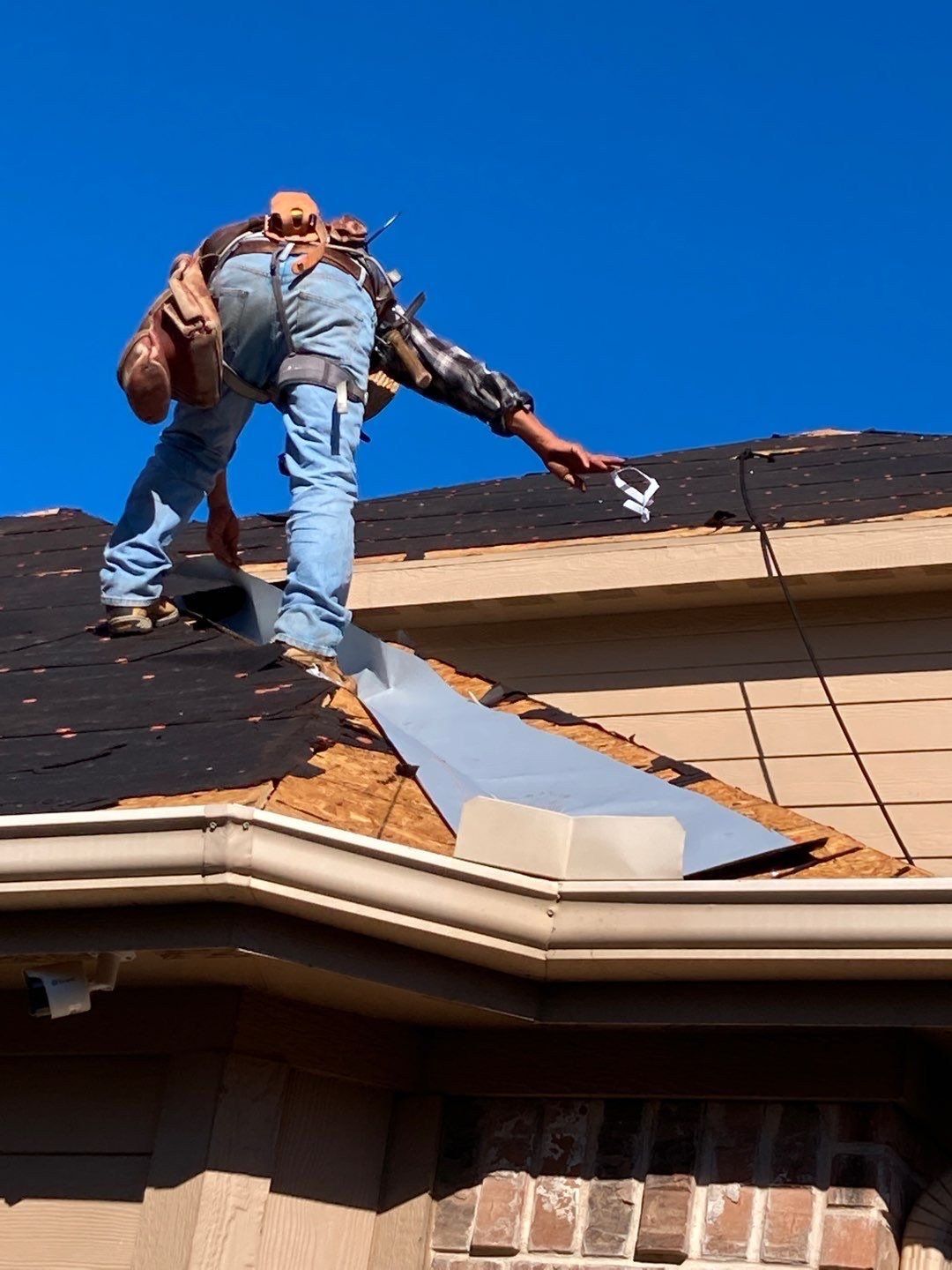 Worker repairing roof shingles beside a silver flashing strip on a house roof under a blue sky