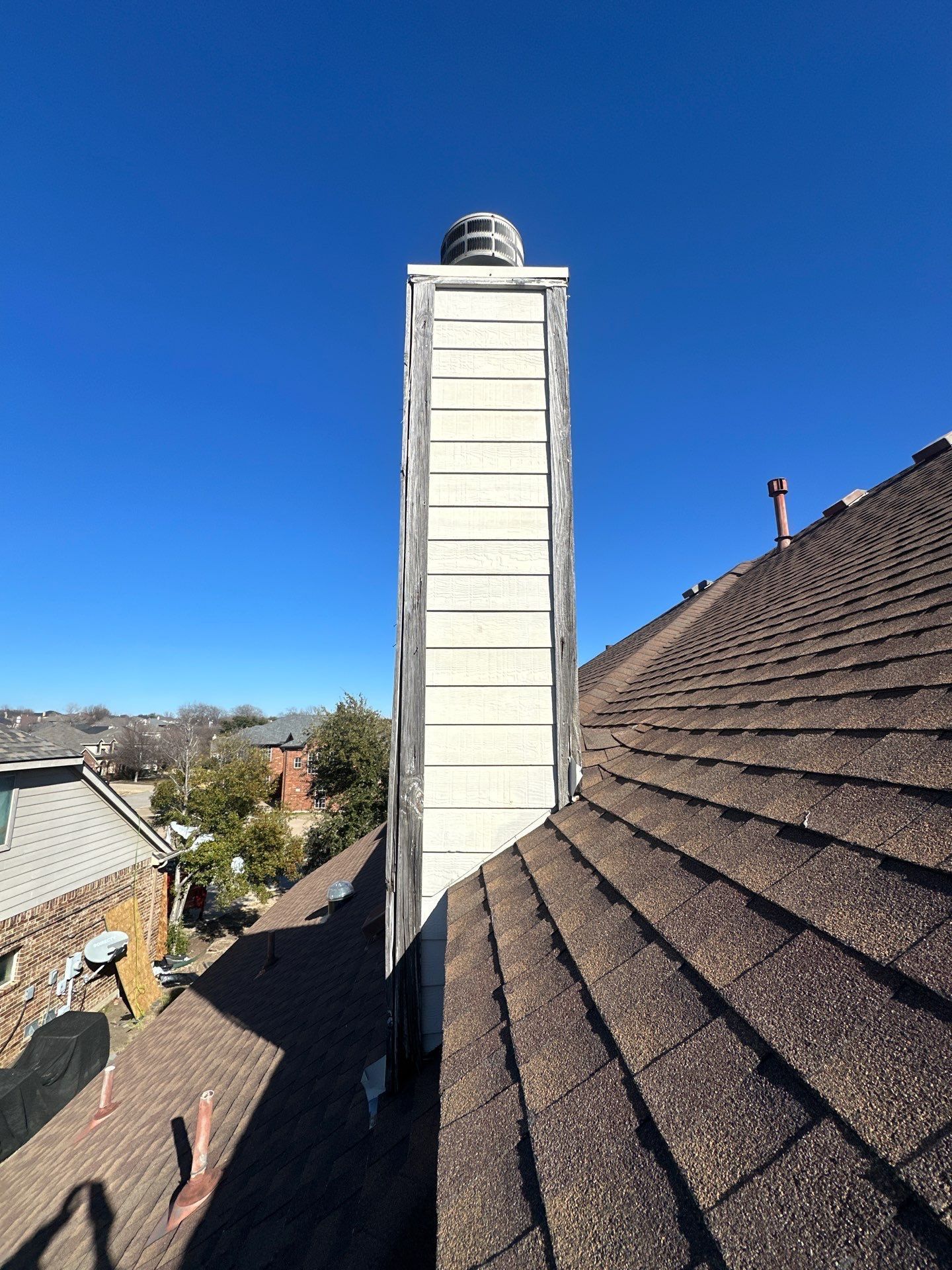 Rooftop view of a tall white chimney beside brown shingle roofs under a clear blue sky