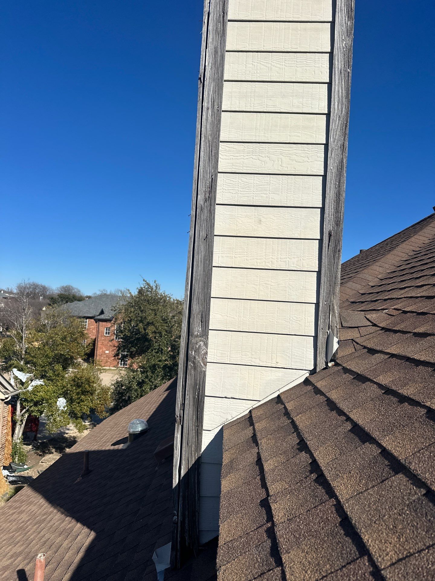 Rooftop chimney with beige siding beside dark shingled roofs under a clear blue sky.