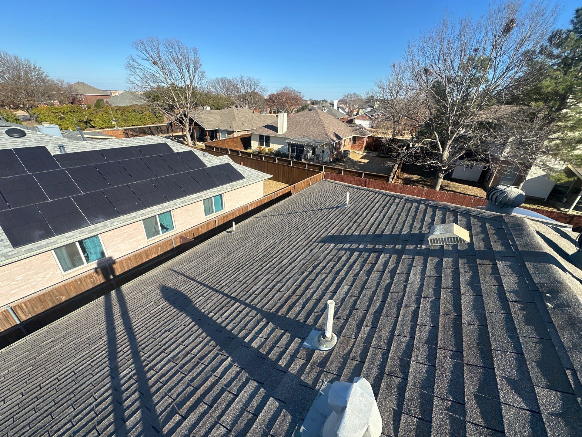 Rooftop with gray shingles, skylights, and solar panels overlooking suburban houses under a clear blue sky