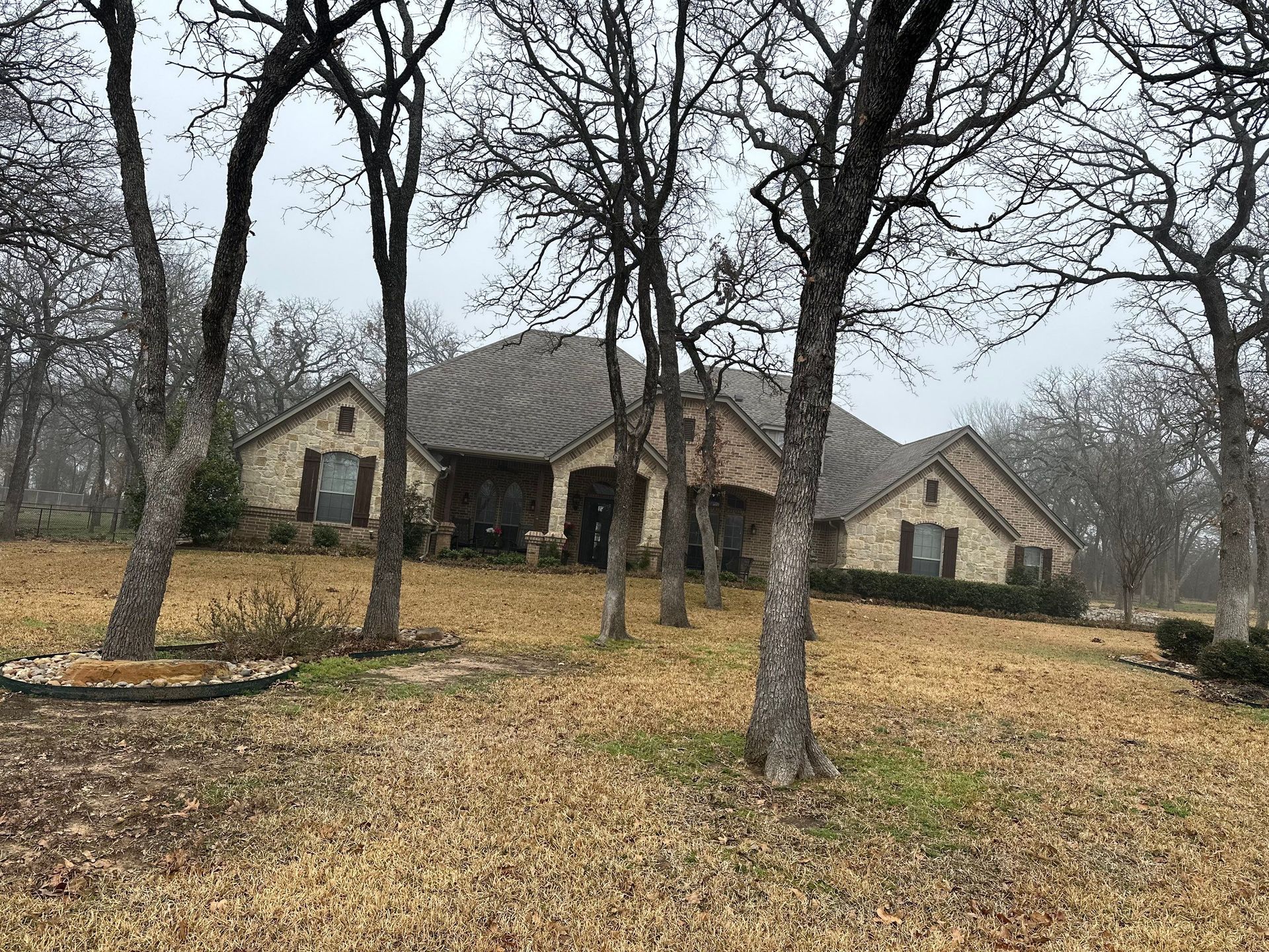Wooded yard with leaf-covered ground and a tan house under bare trees on an overcast day