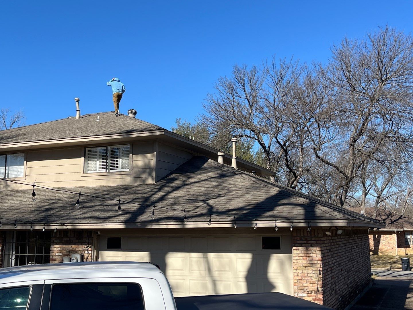 Suburban house with beige siding, dark roof, bare trees, and a white vehicle parked in front.