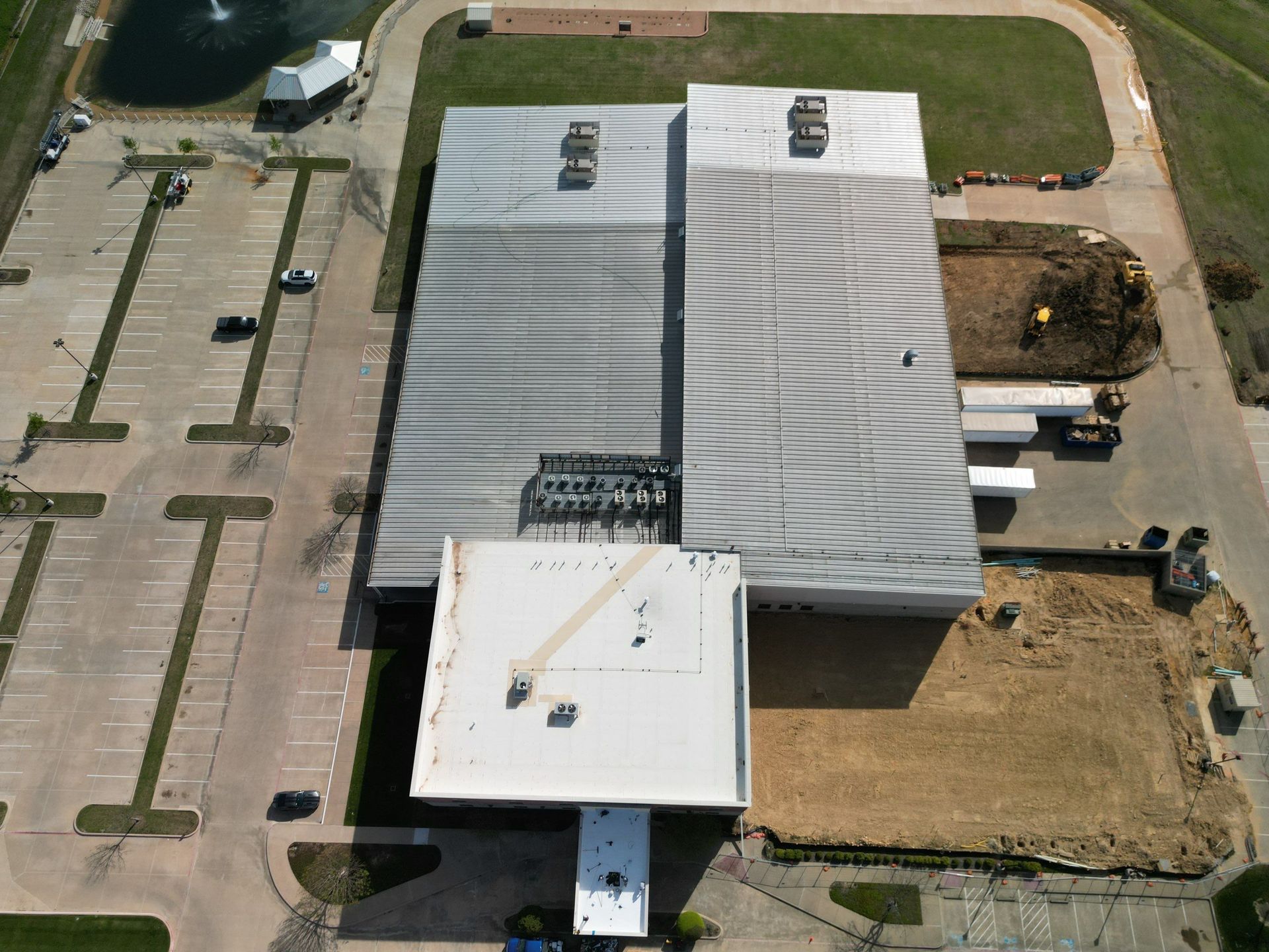 Aerial view of a large industrial building with a white roof, parking lot, and surrounding dirt lot.