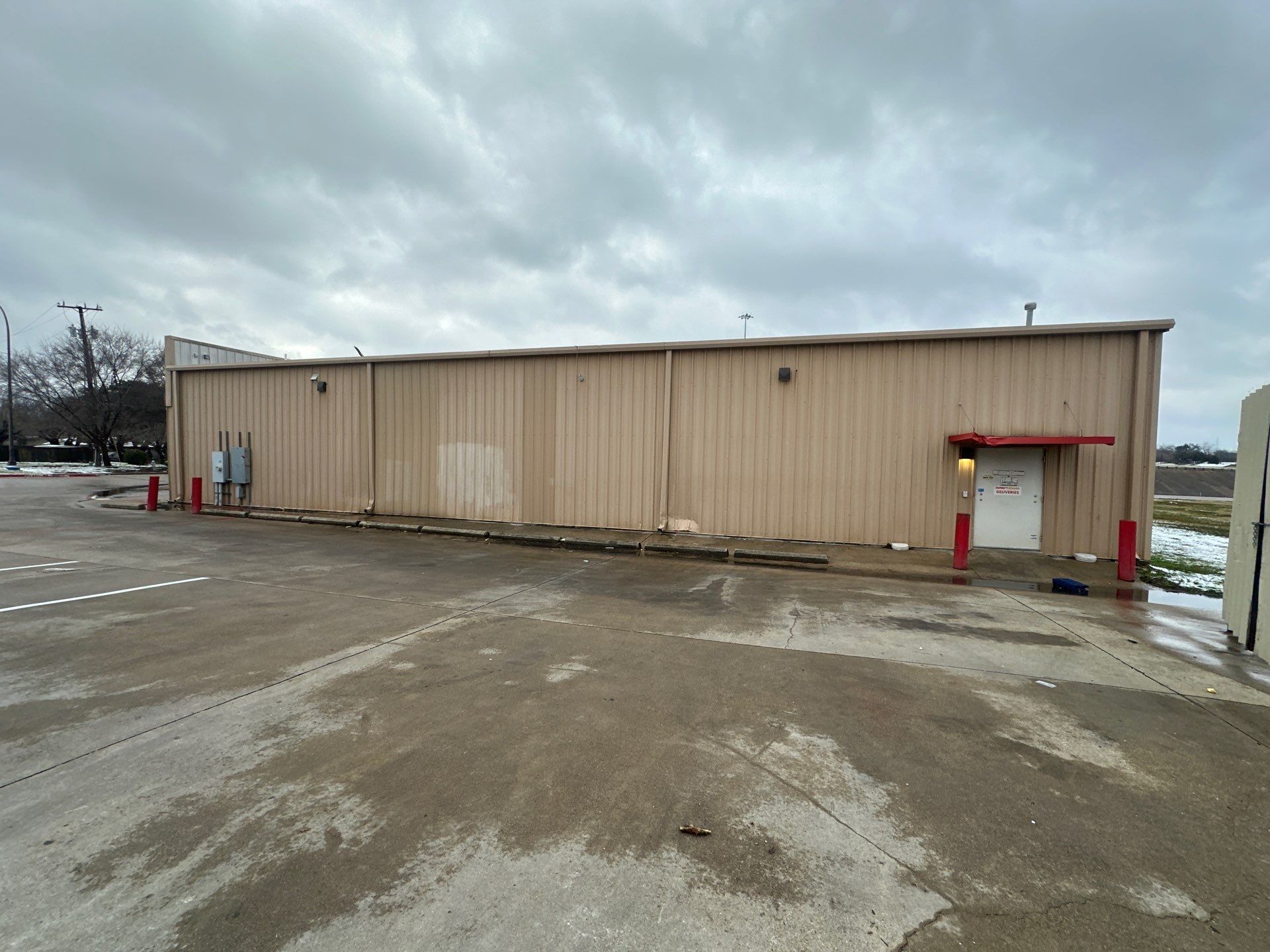 Tan beige utility building with a red awning on a wet, muddy parking lot under a cloudy sky