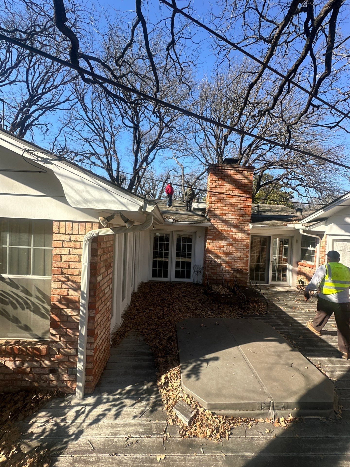 Person in a yellow safety vest walking by a brick house with a chimney and leaf-covered front steps.