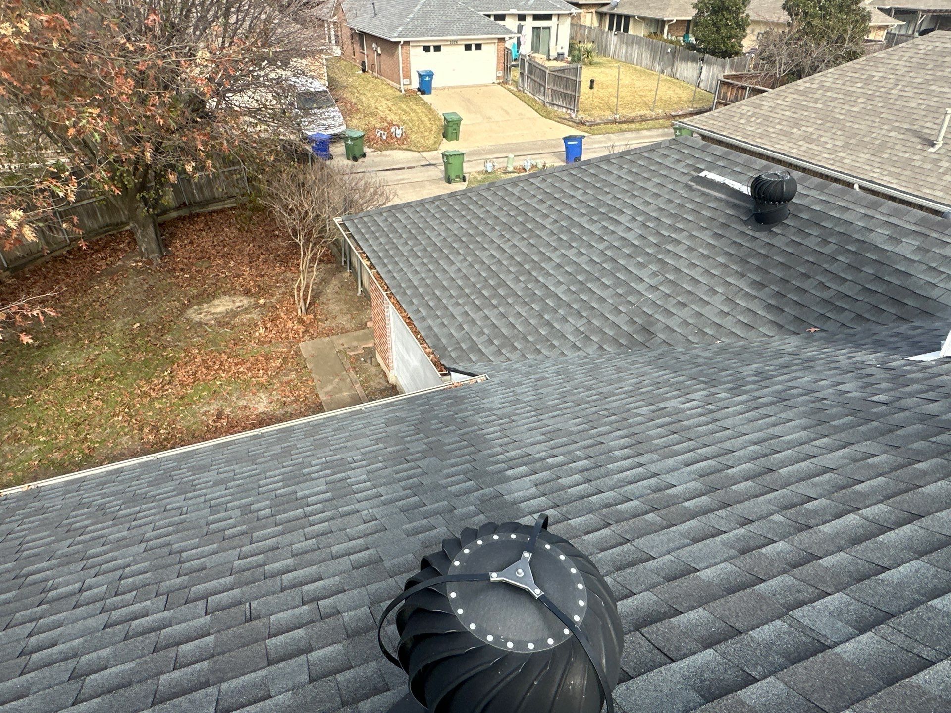 Rooftops with gray shingles and a black vent, overlooking a yard with trees and a walkway