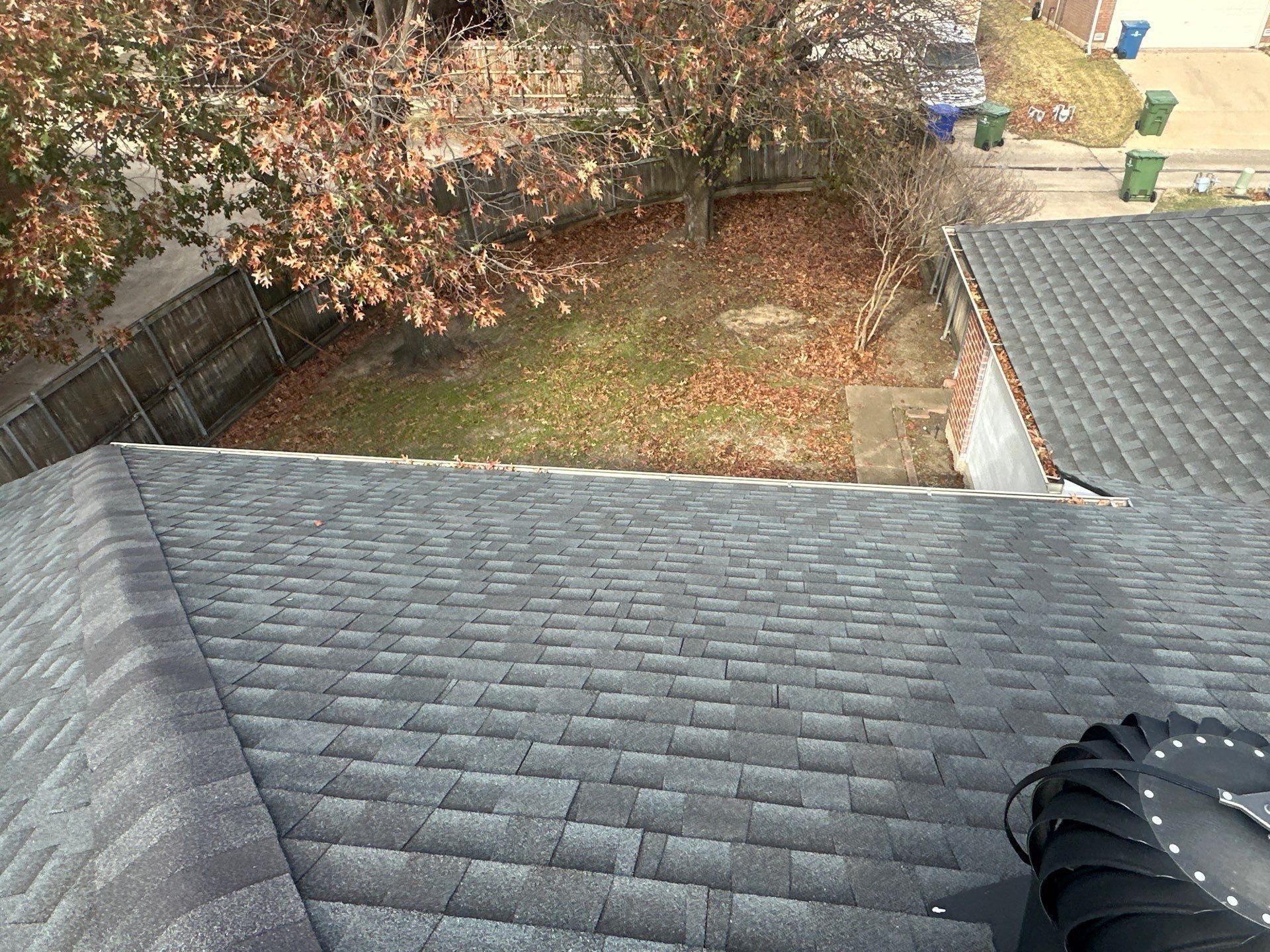 View from a shingled rooftop overlooking a fenced backyard with autumn trees and a patio below