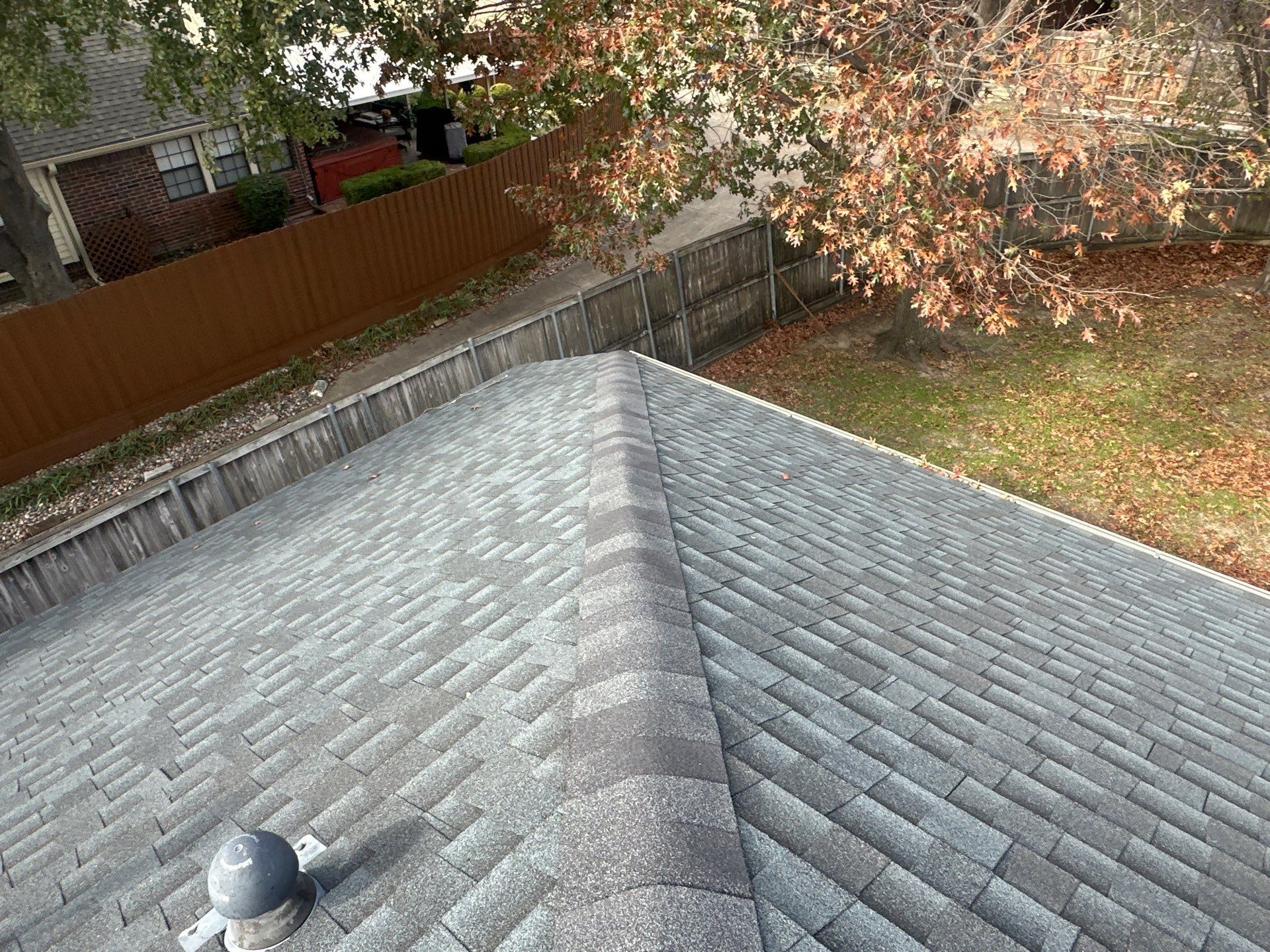 Gray shingled roof with a vent, bordered by a wooden fence and autumn trees in the yard