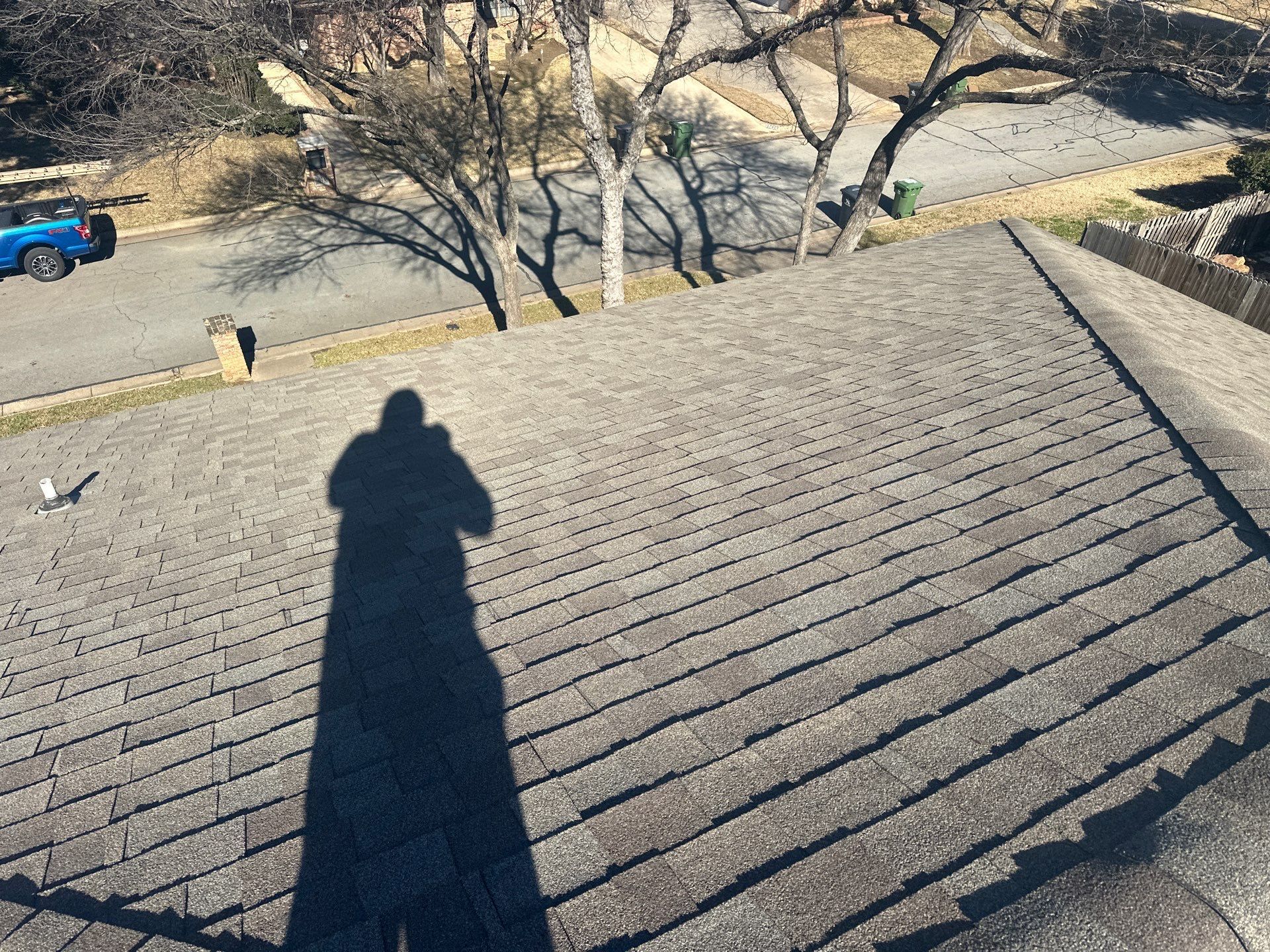 Shadow of a person on a sloped shingled roof, with trees and a road below.