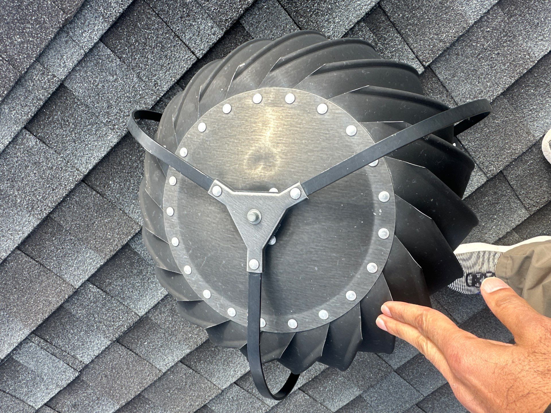 Close-up of a roof vent fan with black blades and a hand pointing at it on gray shingles