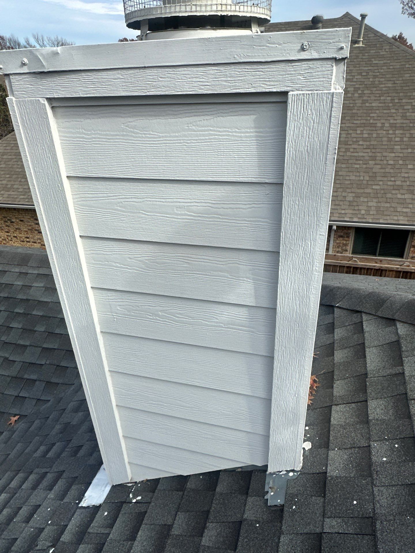 Gray rooftop chimney chase with horizontal siding and a metal cap, on a shingled roof.
