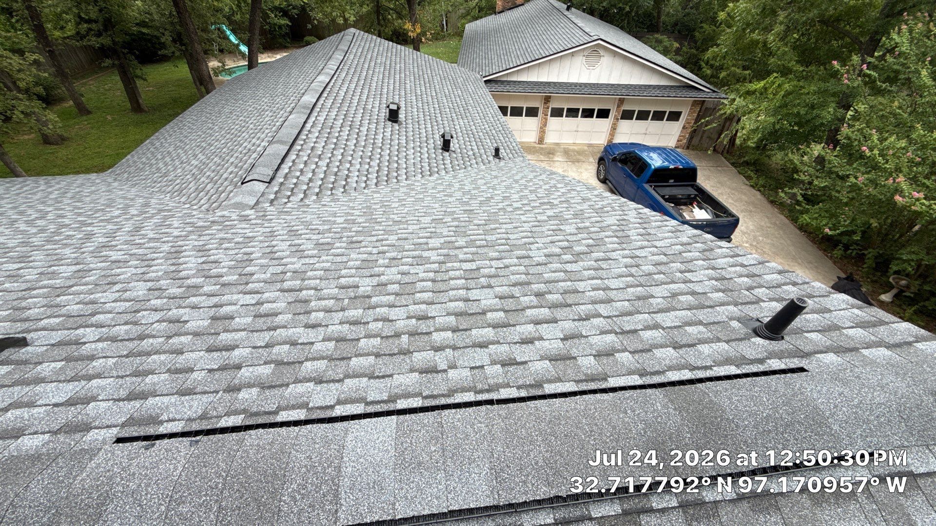 Roof covered in gray shingles beside a garage, with a blue car parked in the driveway under trees.