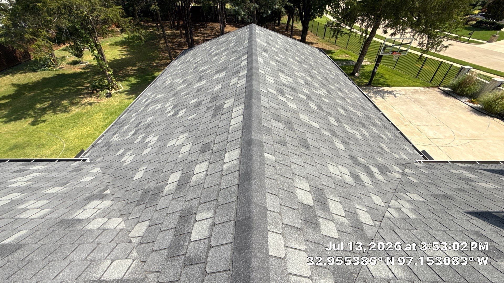 Gray shingled roof ridge in a sunny wooded residential area with lawns and a paved driveway