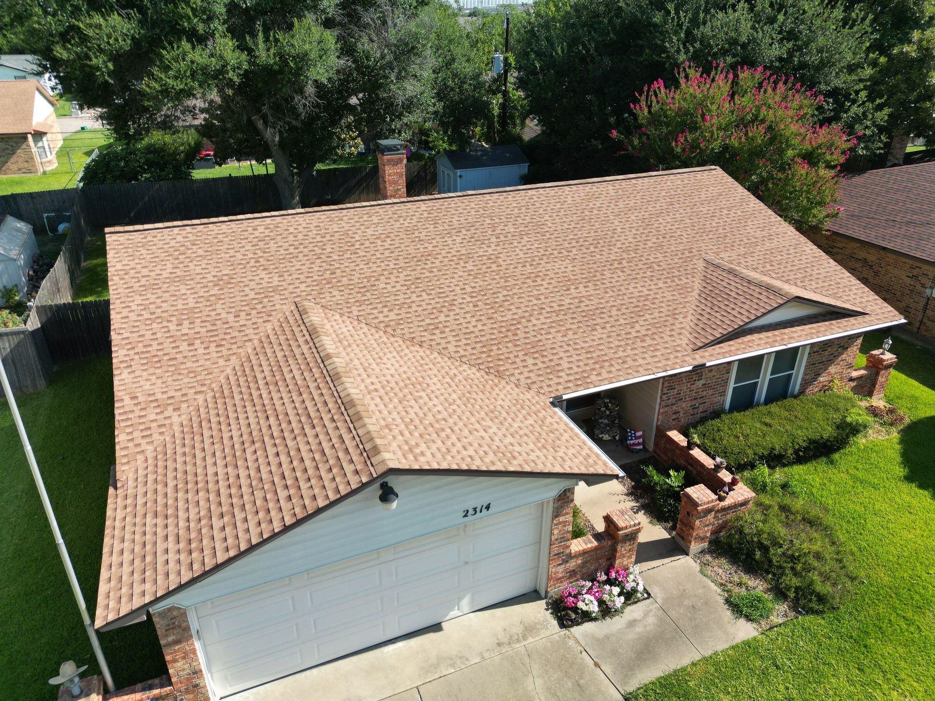 Aerial view of a tan-roofed suburban house with a garage, front porch, and green yard.