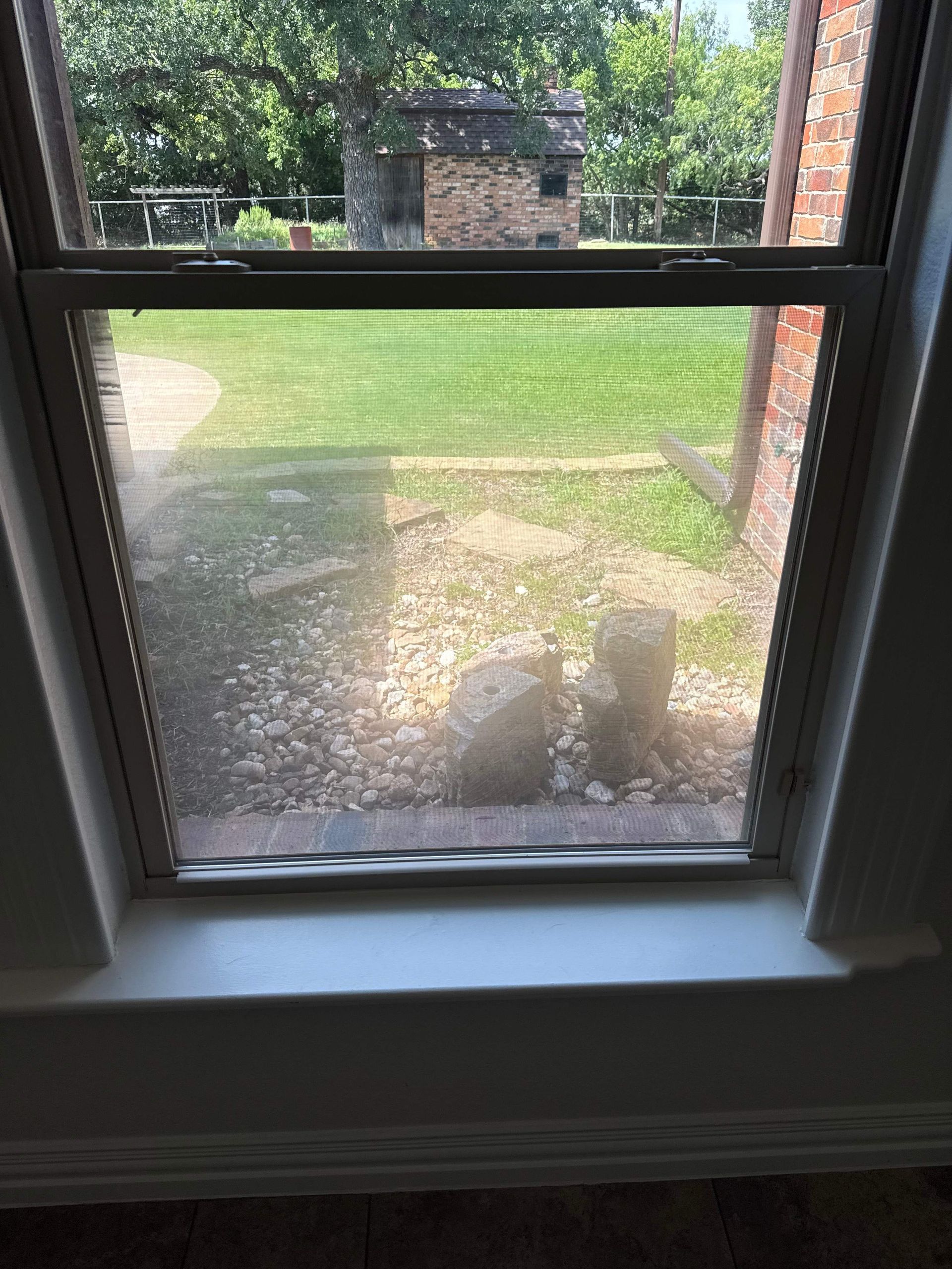 View through a window to a backyard with a brick wall, green lawn, and a small cat on the patio.