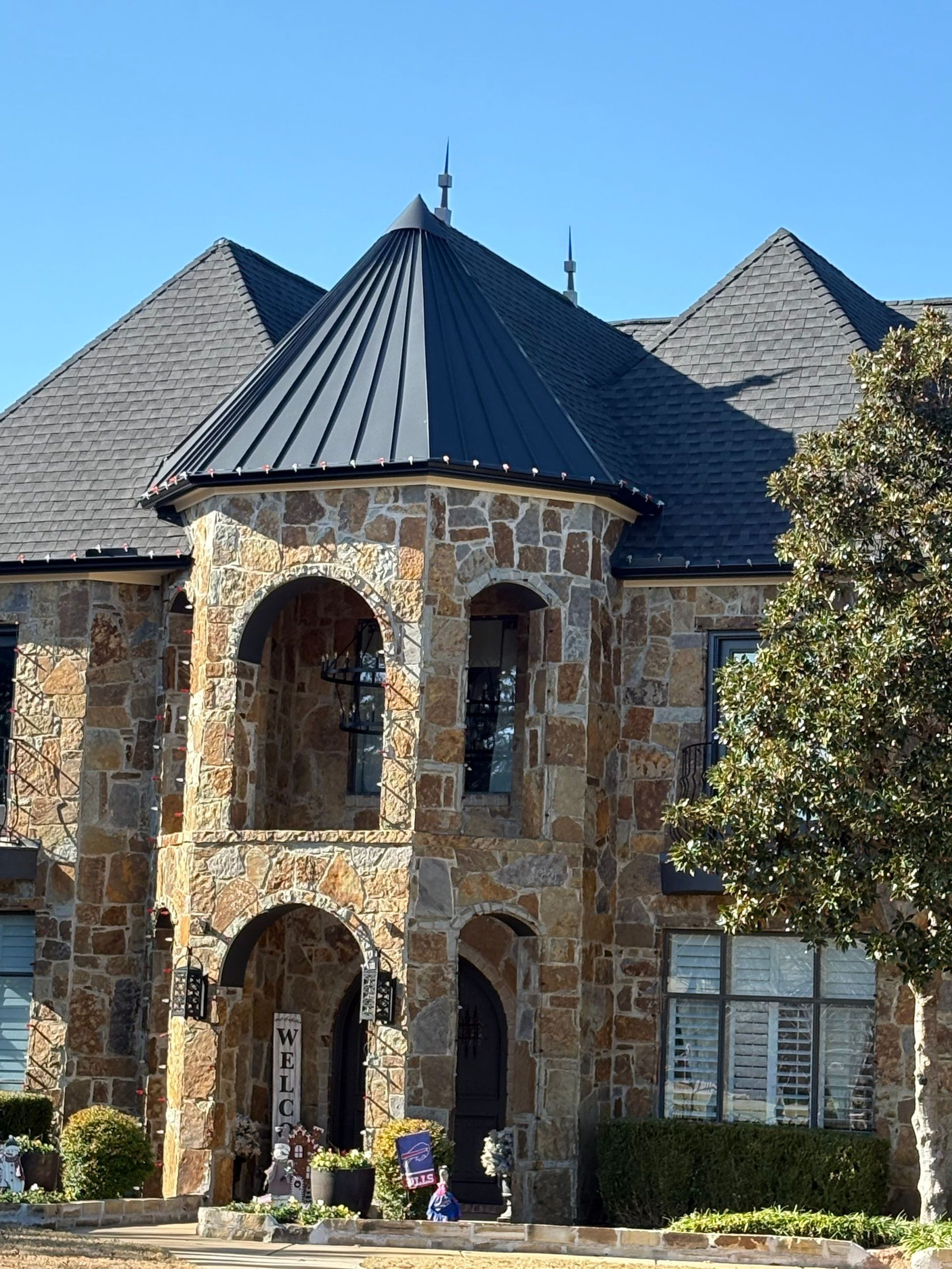 Stone turret entrance with arched windows and dark roof on a large brick house, with flowers below.
