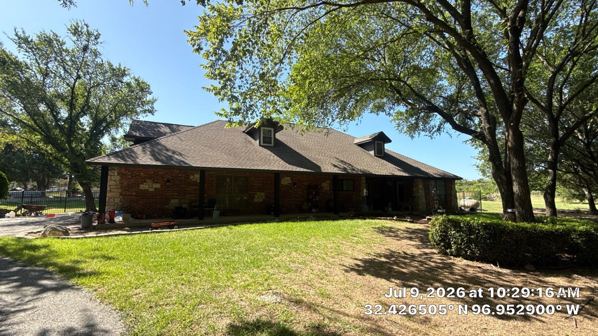 Single-story brick house with a metal roof and trees in a sunny yard