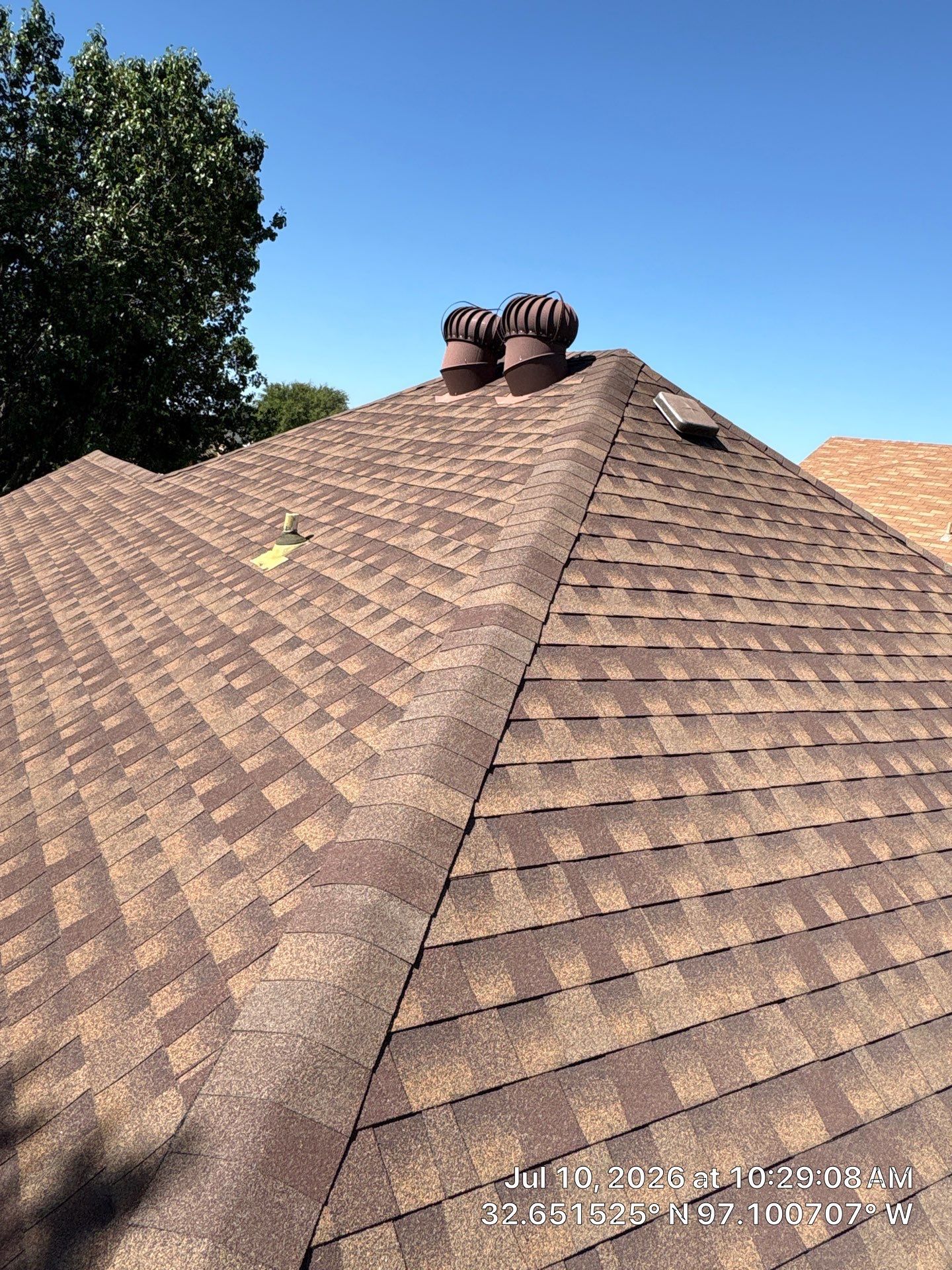 Brown shingle roof with two vent stacks, viewed from above under a clear blue sky
