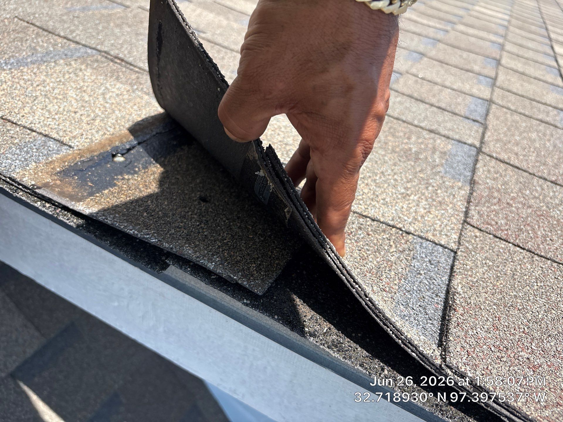 Hand inspecting a lifted roof shingle on a sloped shingle roof, with a nearby skylight vent.