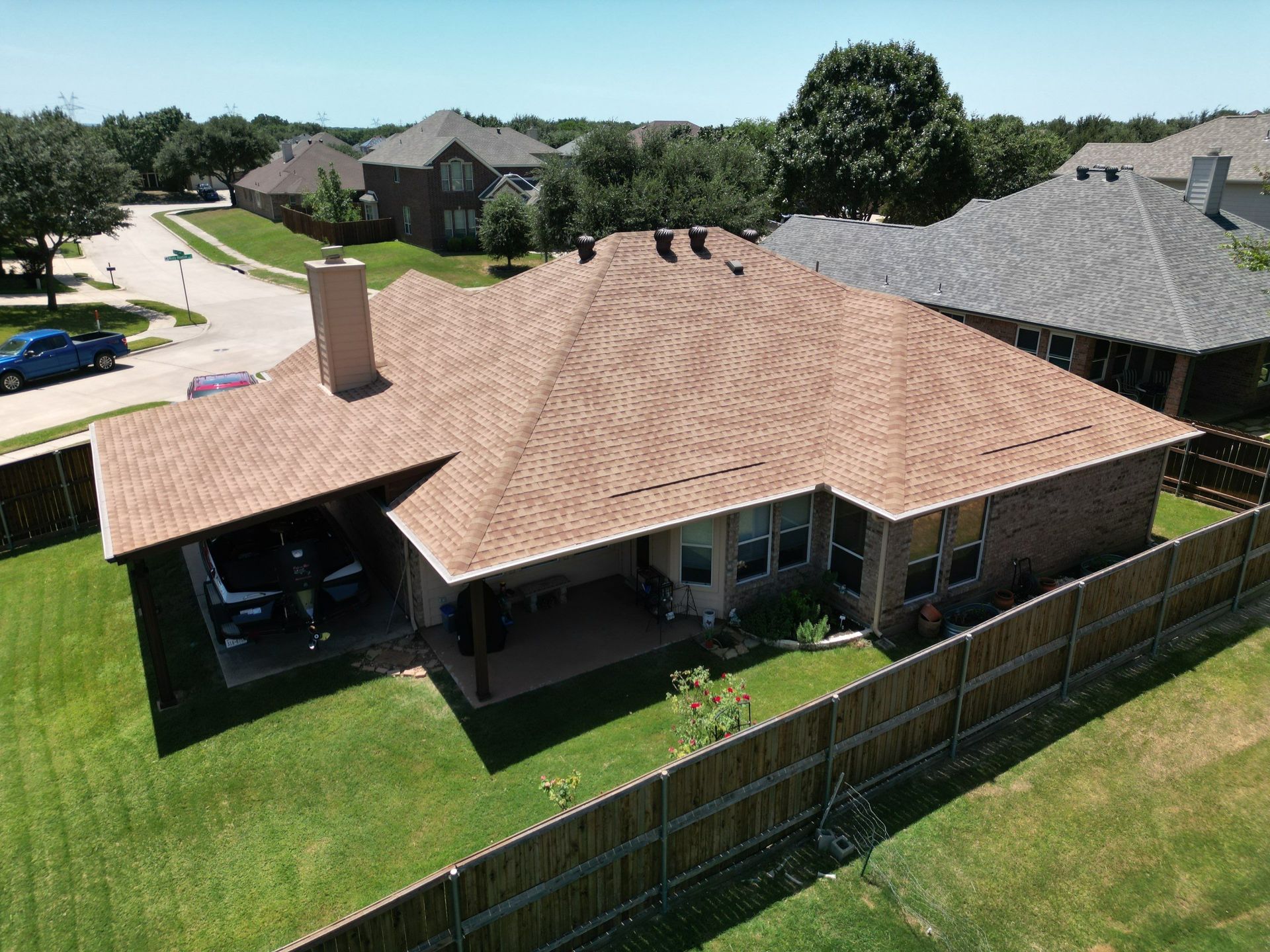Aerial view of a suburban house with a tan roof, fenced backyard, and neighboring homes on a sunny day
