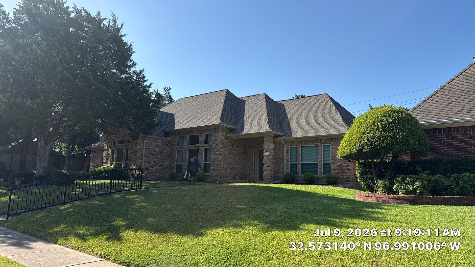 Brick suburban house with a gray roof, green lawn, and trees under a bright blue sky