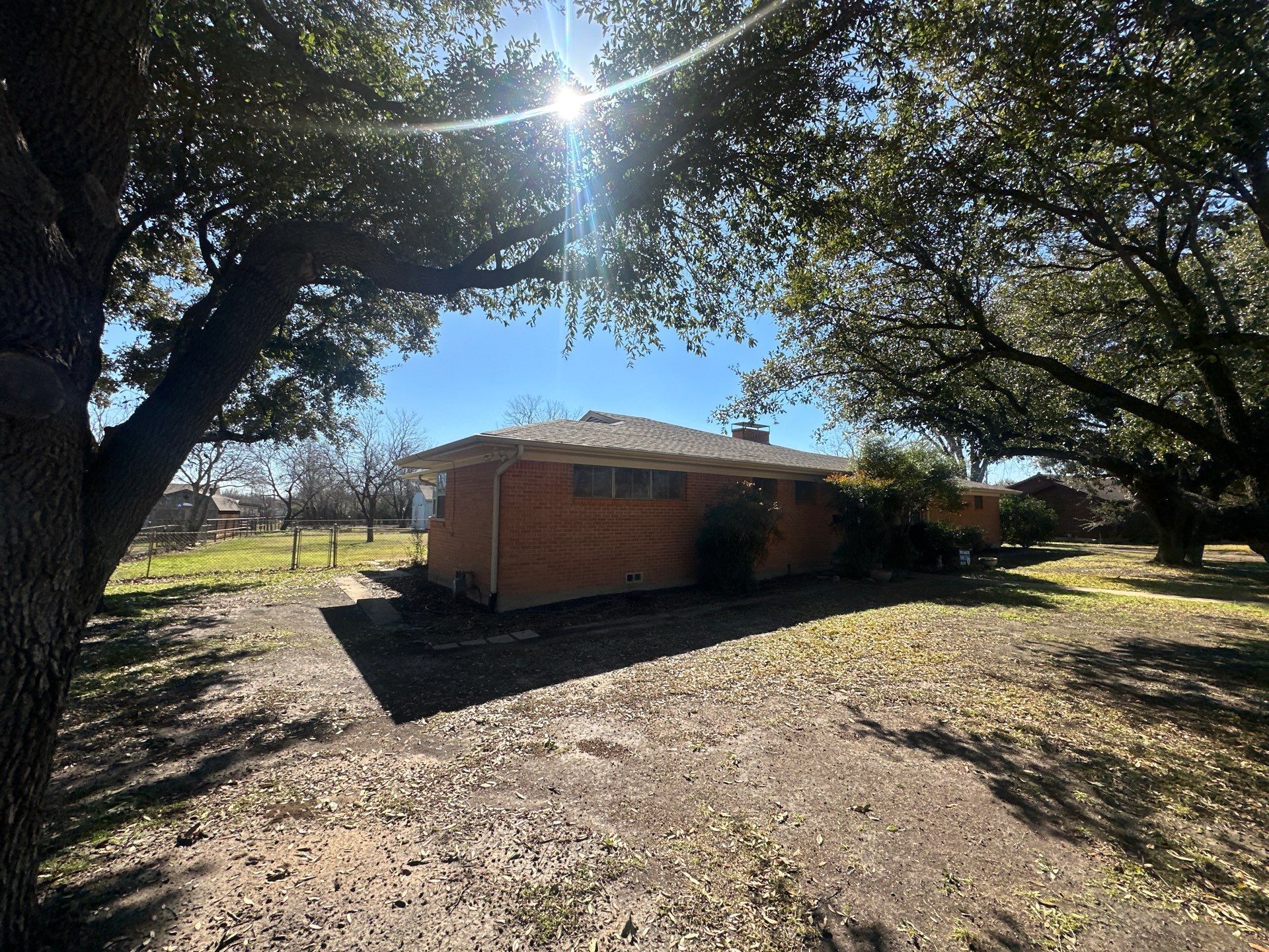 Brick house shaded by large trees on a sunny dirt yard with open field nearby