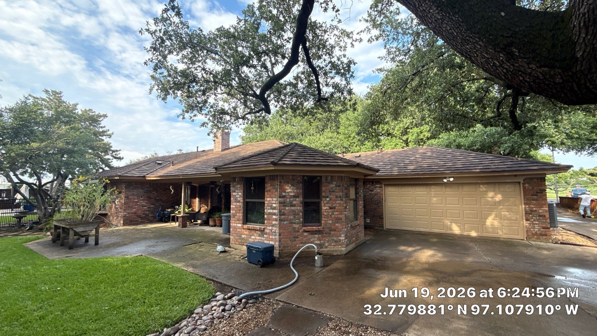 Single-story brick house with a front porch, large trees, and a wet driveway under a cloudy sky.
