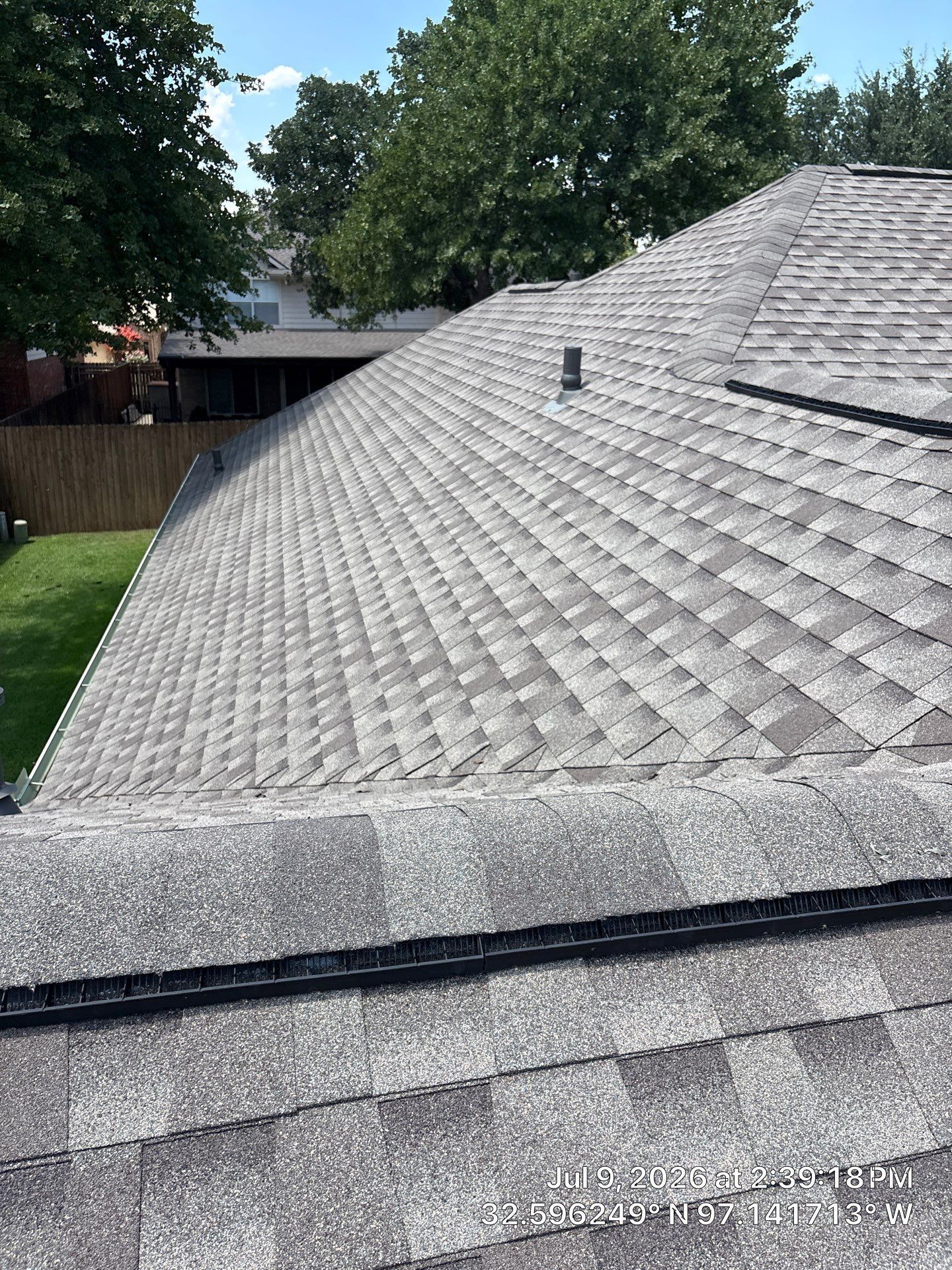 Gray shingle roof on a house, viewed from above with trees and a fence in the background