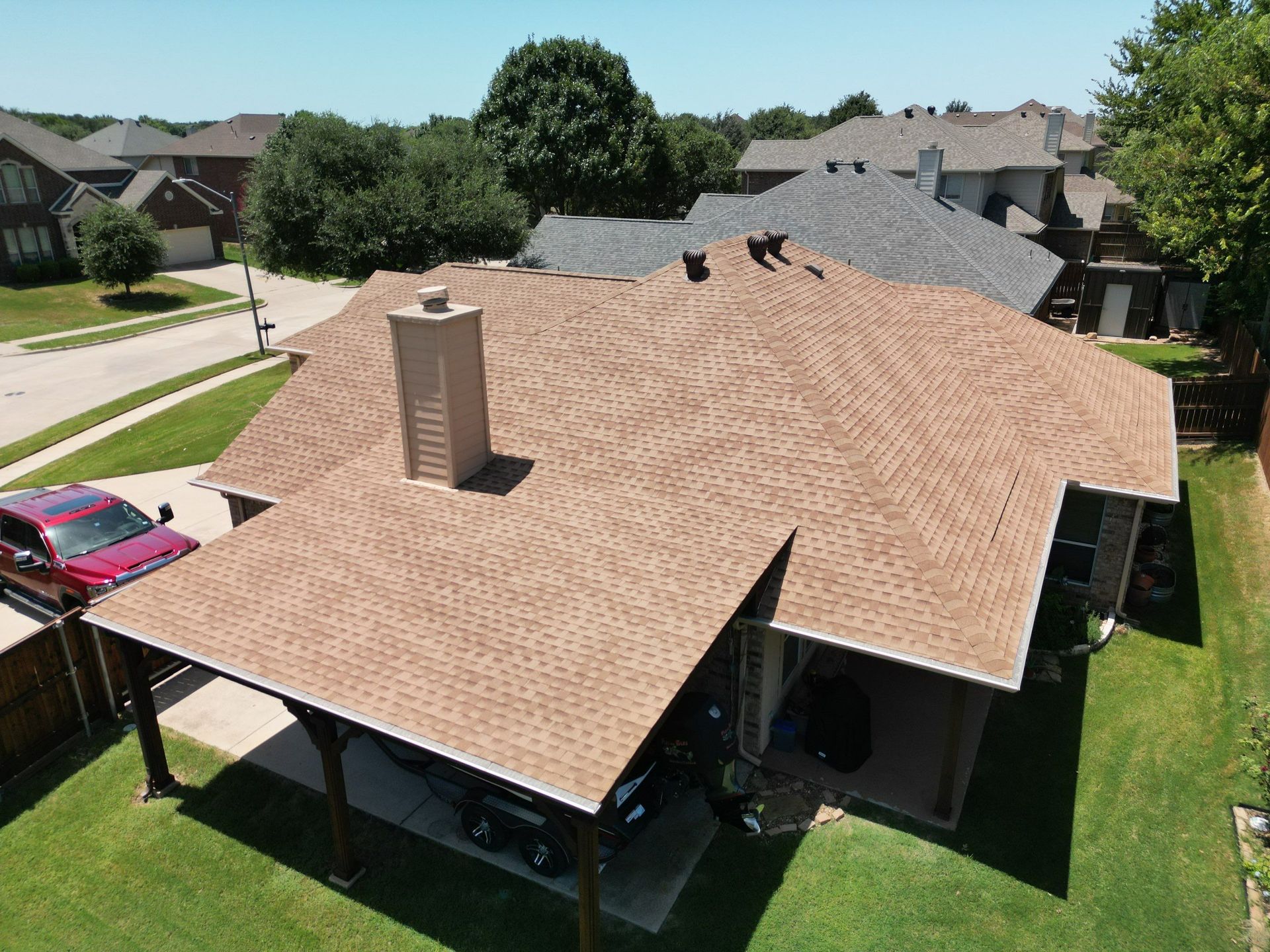 Aerial view of a suburban house with a tan shingle roof and a covered patio, surrounded by green lawn.
