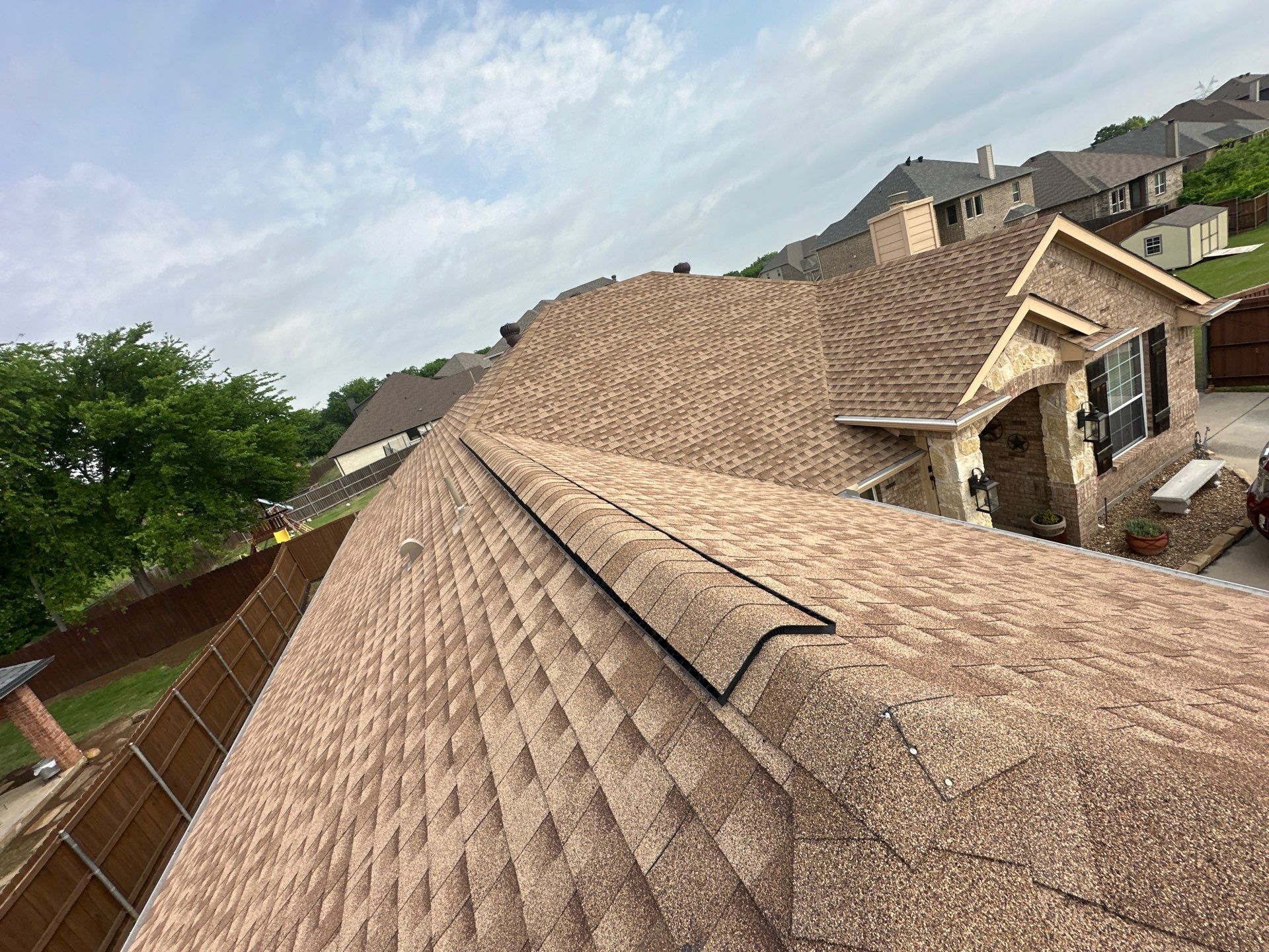 Roofing under construction on a tan suburban house, viewed from above beside a fenced yard.