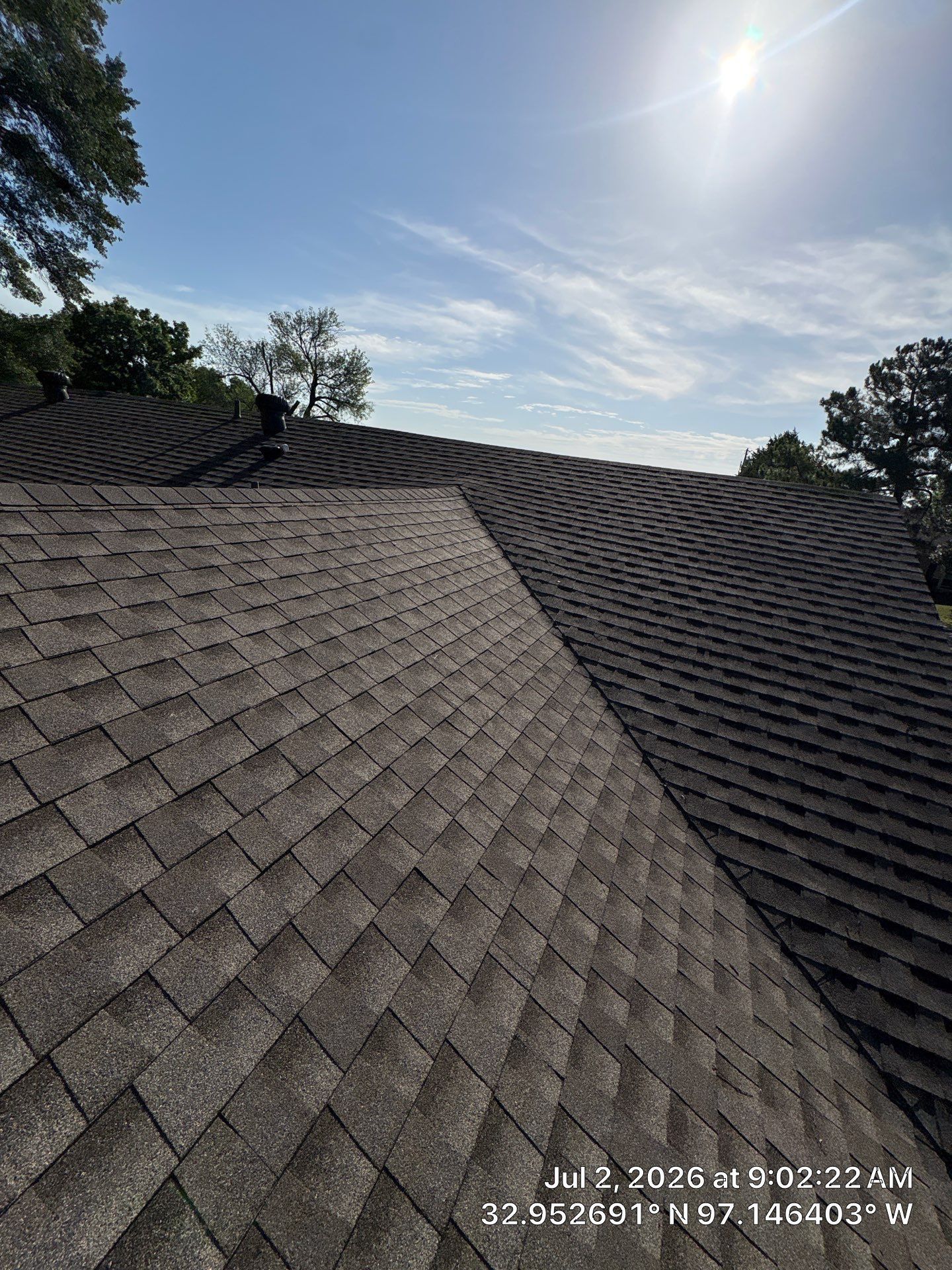 Sunlit shingled roof with dark gray sections and blue sky above trees