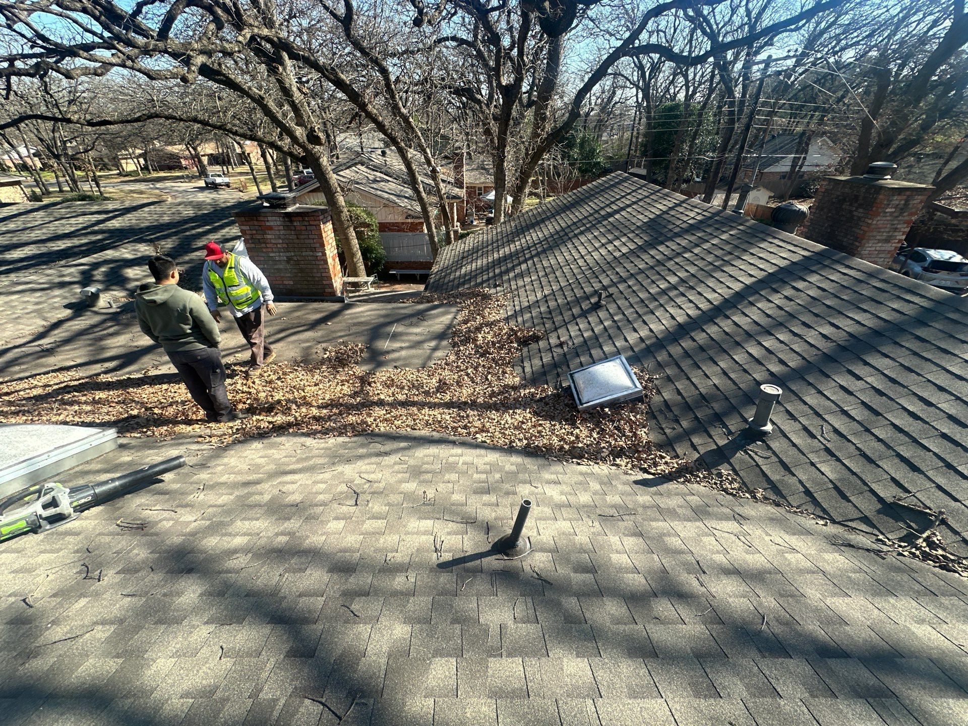 Workers on a shingled roof clearing debris near trees and a small shed