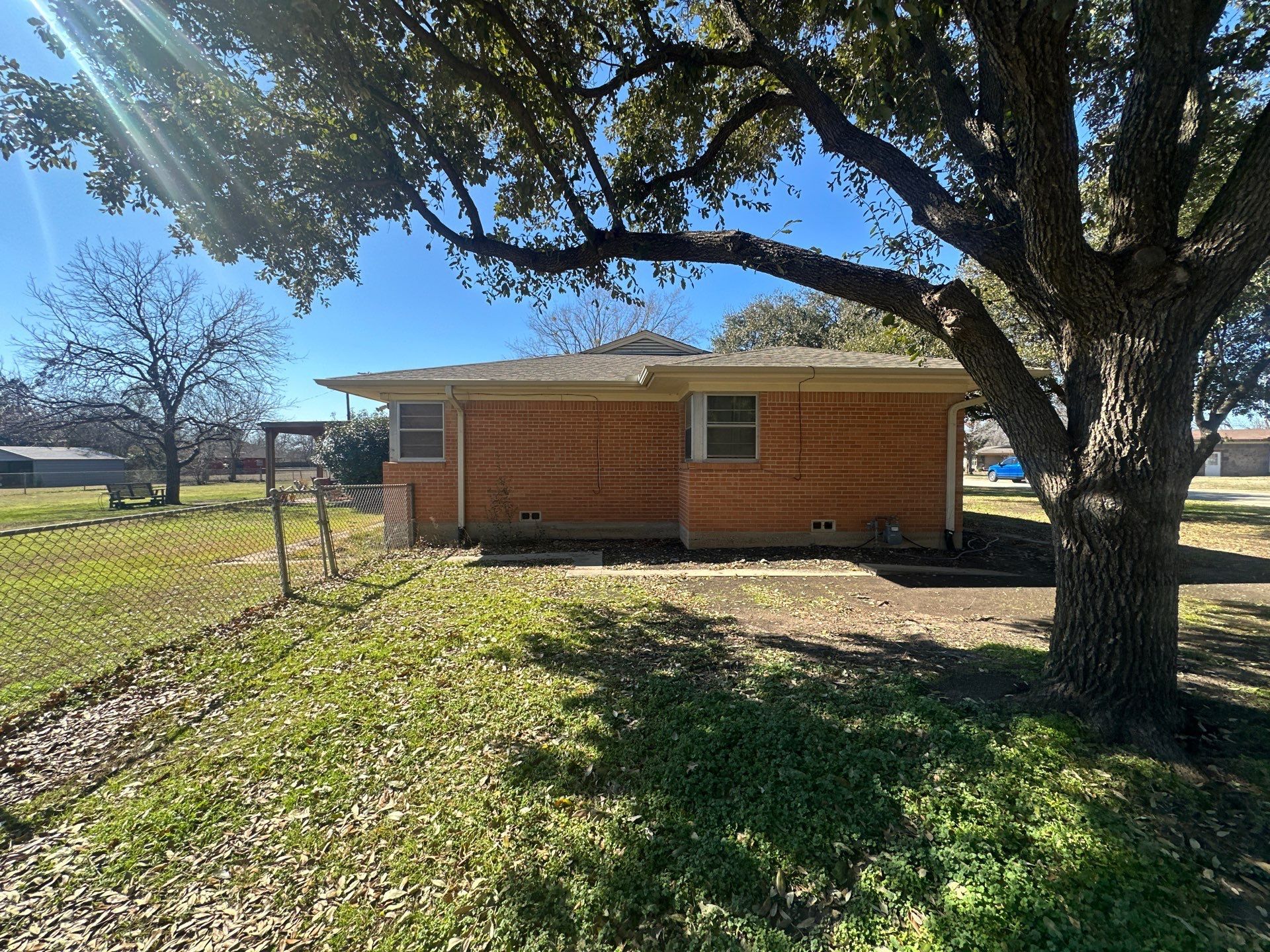 Small brick house with a large tree casting shade over a grassy yard on a sunny day